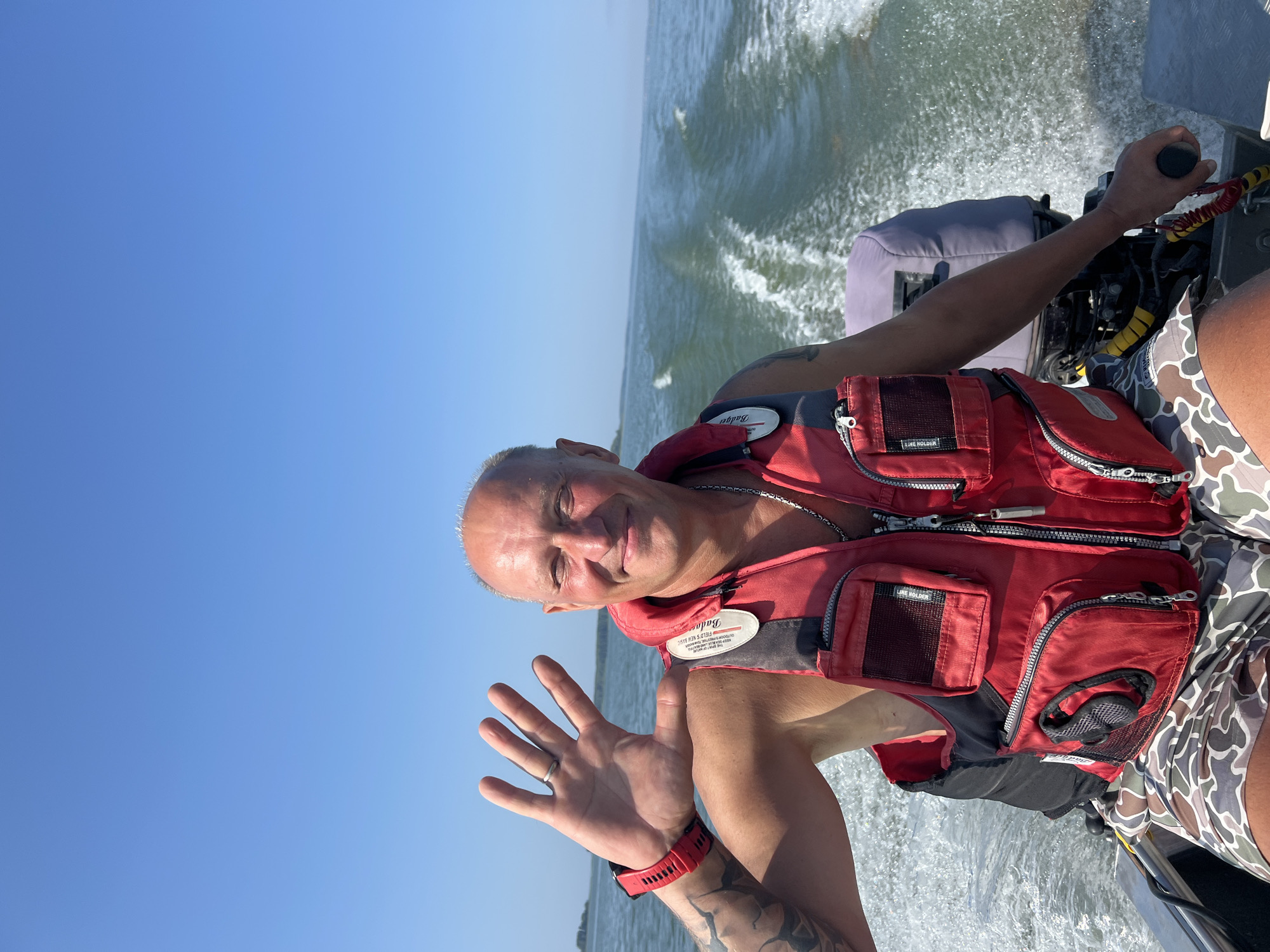 A man is on a boat, waving to the camera. He is wearing a red life jacket and camouflage shorts. The boat is moving through the water, leaving a wake behind it. The sky is clear and blue, and there are trees and buildings in the distance. The location is the city of Beregovoe, Russia.