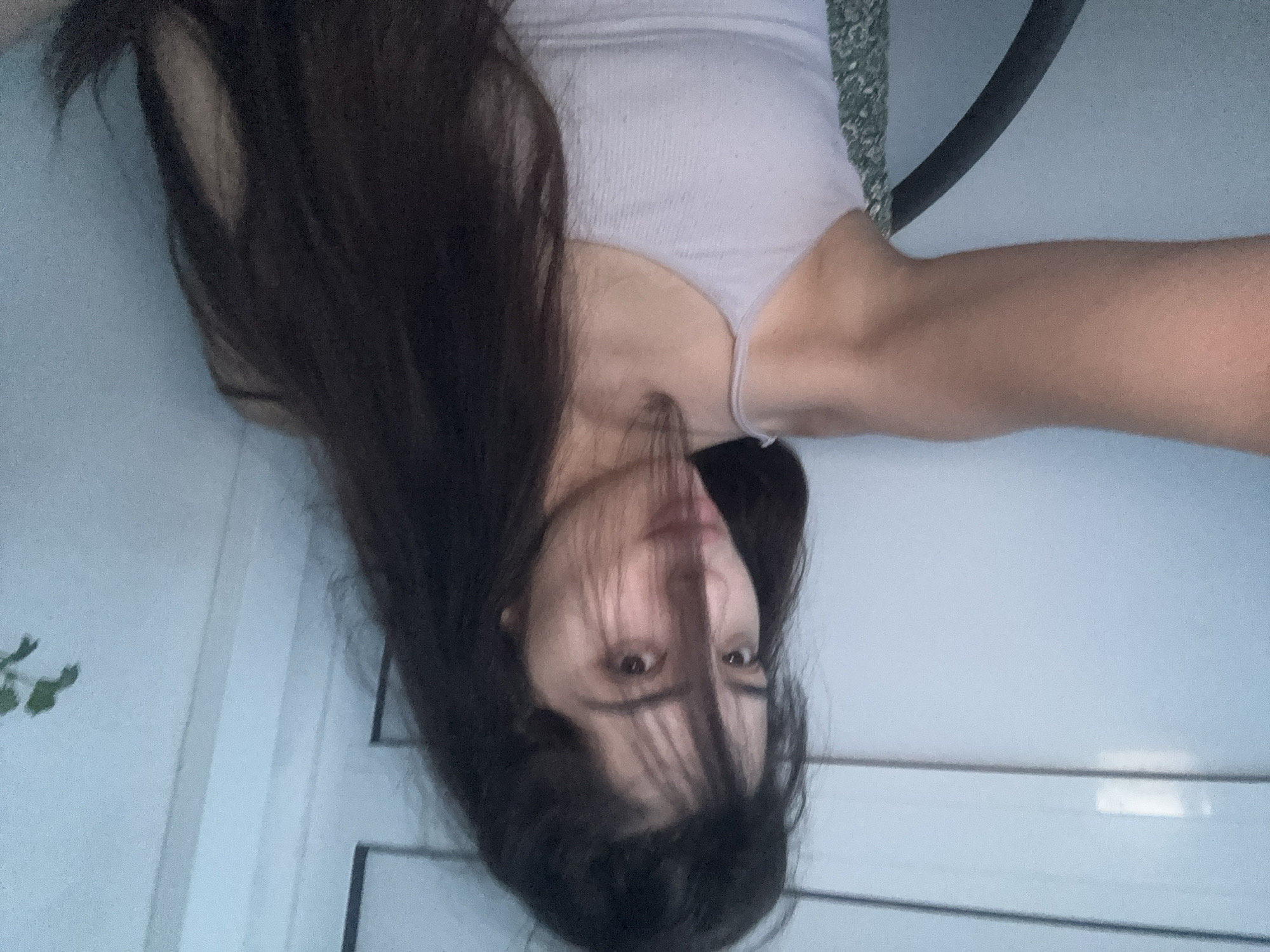 The image displays a young woman with long, dark brown hair, appearing upside down due to the photograph's 90-degree clockwise rotation. She is wearing a light-colored, ribbed tank top, possibly pale pink or lavender, with thin straps. Her face, partially covered by strands of her hair, looks directly towards the camera with a neutral expression. Her left arm is extended and visible towards the right side of the frame.
The setting appears to be indoors, against a predominantly plain, light-colored background, likely a wall. On the left side of the frame, subtle vertical lines suggest a doorframe or architectural molding, and a small amount of green foliage is visible in the bottom left corner, possibly from a houseplant or a view through a window. Behind her left shoulder, a textured, mottled greenish-grey surface is visible, which could be part of a blanket or cushion. A dark, curved object, possibly part of furniture or a cable, is also present near her right shoulder. The lighting is soft and even, suggesting an indoor environment or diffused natural light. No visible text is present in the image.