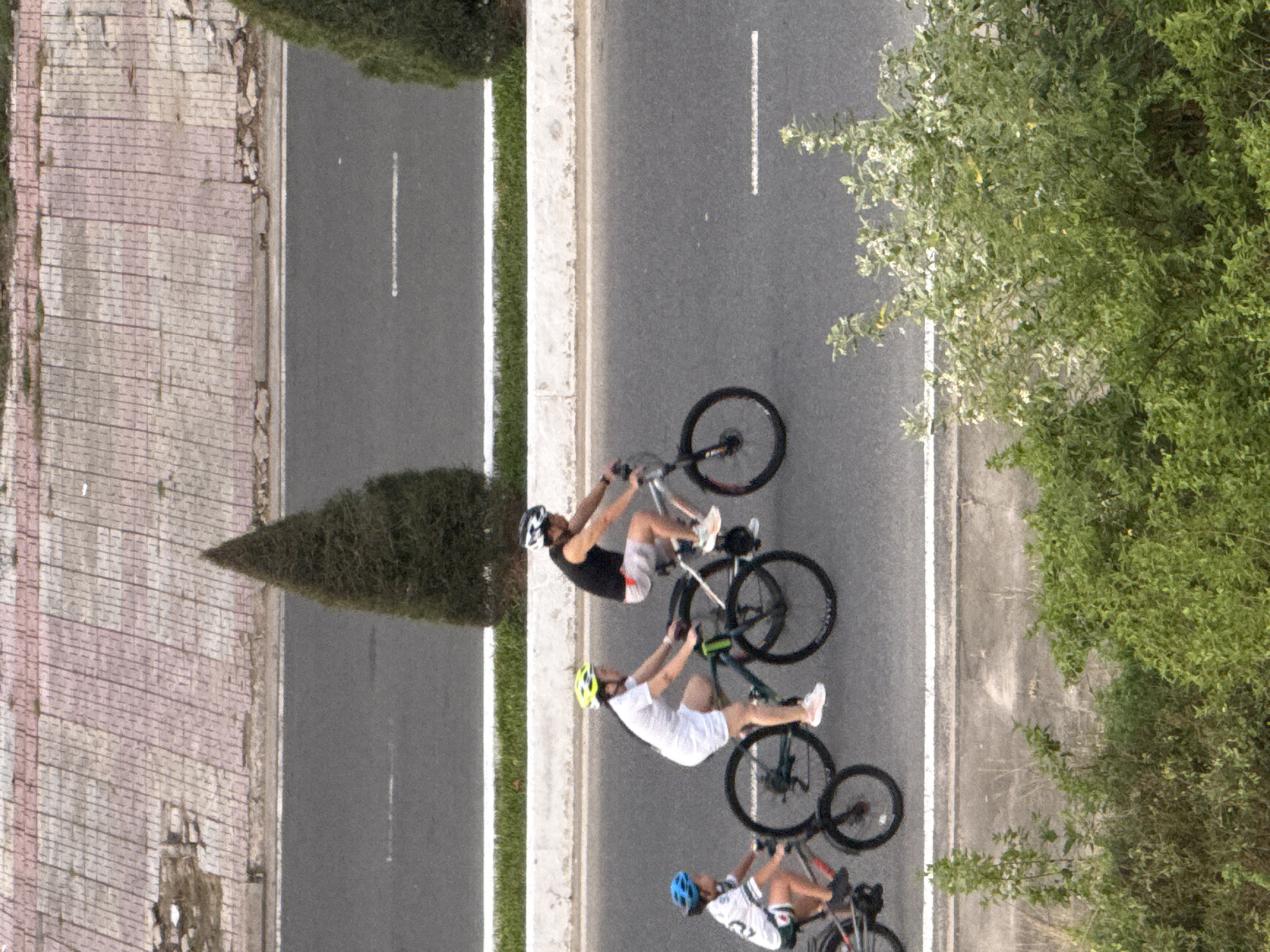Three individuals are cycling on a paved road. The person on the left is a child wearing a white and green shirt and a blue helmet, riding a dark bicycle. In the middle, an adult in a white shirt and shorts with a yellow helmet is riding a dark bicycle. The person on the right is an adult wearing a black tank top and light-colored shorts with a white helmet, riding a lighter colored bicycle. They are all moving in the same direction. The road is flanked by a concrete barrier and grassy areas. In the background, a tiled walkway with some debris is visible, as are two conical trees. The scene appears to be outdoors during daylight, possibly in Bac Nha Trang Ward, Vietnam, given the context.