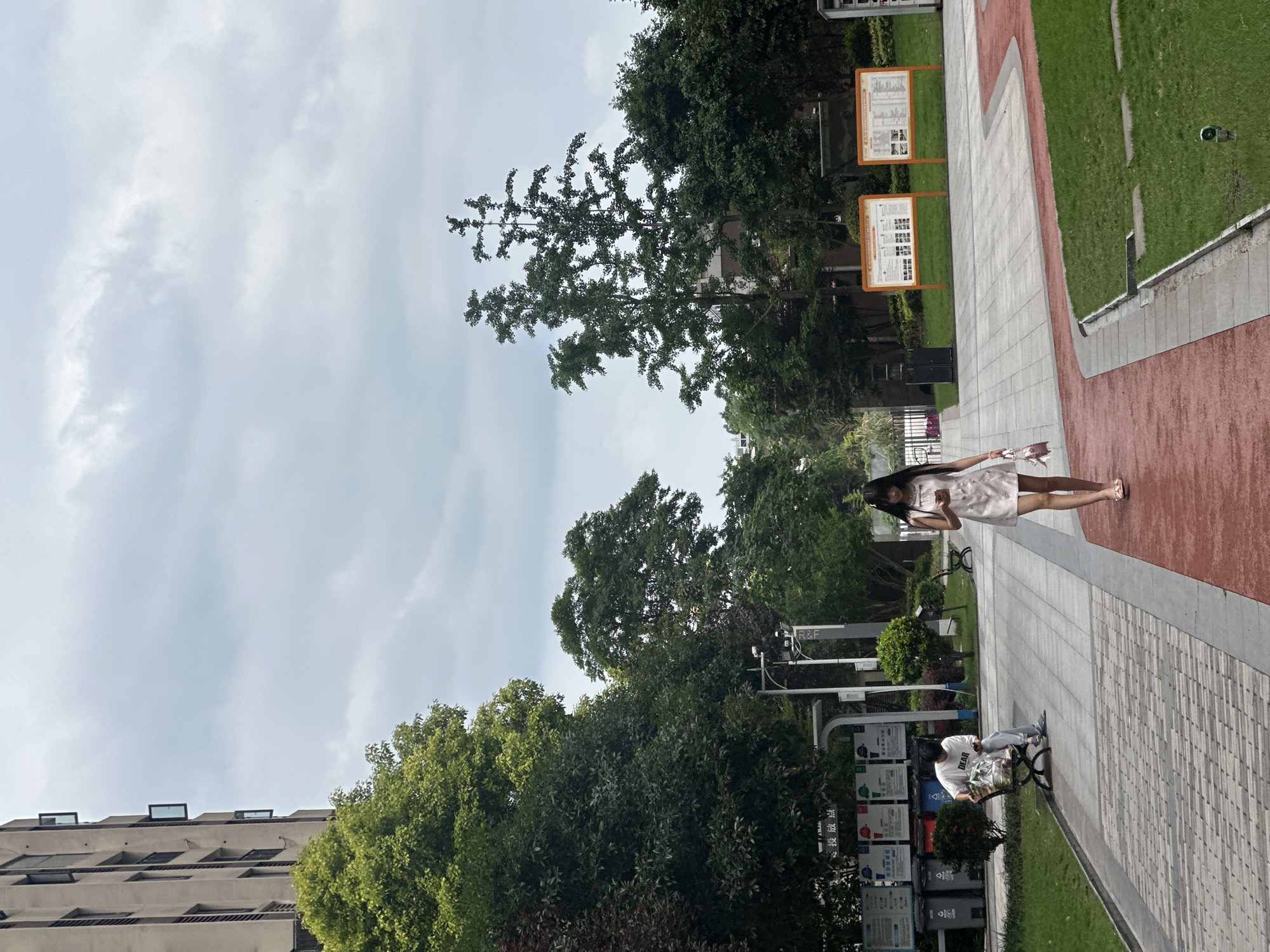 The image shows a partially obscured exterior view in the Chongqing High-tech Zone, China. A woman in a short, light-colored dress and sandals walks along a pathway in a park-like setting. A man is seated on a bench near trash receptacles and signage. The scene features trees, grass, paved areas, and informational displays. A building is visible in the background. The sky is partly cloudy.