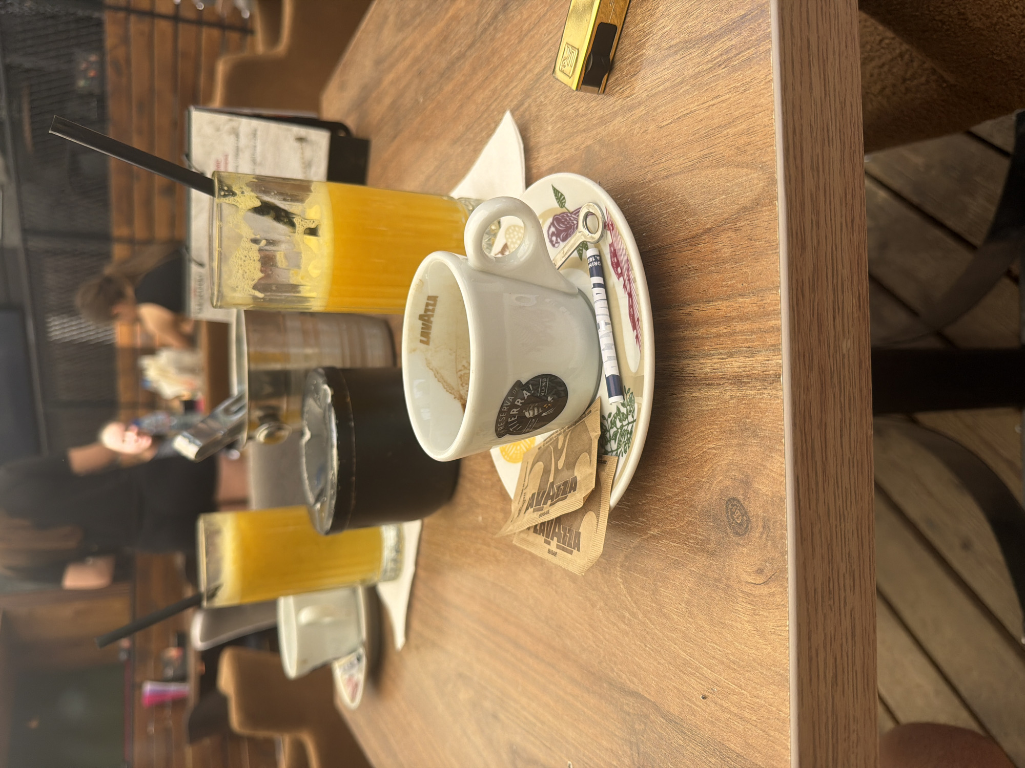The image depicts a detailed indoor or covered outdoor café or restaurant scene in Shumen, Bulgaria. The central focus is a light brown wooden tabletop exhibiting distinct grain patterns and a visible knot in the wood.
In the foreground, a white ceramic espresso cup, with "LAVAZZA" printed on its interior rim and "PRESERVA DE TIERRA! BLEND" alongside a bird/owl logo on its exterior, contains residual dark brown coffee stains, indicating it has been used. This cup rests on a white ceramic saucer adorned with a green foliage and a red/pink bird motif. On the saucer, two brown paper packets, one partially obscured but visible with "LAVAZZA" branding, are placed. A small white-handled stirring spoon with a silver-colored key-shaped end also lies on the saucer. To the immediate right, a rectangular gold-colored metallic object, likely a lighter or a small tool, is positioned.
Slightly behind the espresso cup and to its left, a black circular ceramic ashtray is present. To the right of the ashtray, a tall, clear glass filled with a vibrant yellow-orange liquid, consistent with orange juice, features a black drinking straw and a foamy head. Further left, another similar tall, clear glass of yellow-orange liquid with a black straw is visible. A white ceramic coffee cup and saucer are partially discernible behind this second juice glass. A metallic bucket, possibly an ice bucket, is also partially visible behind the ashtray. A white rectangular paper napkin is situated beneath the left-most juice glass.
The midground and background feature out-of-focus elements of the café environment. Patrons are seated at other tables; one person with long, dark hair and wearing a dark top is visible from the back on the left, while another person with lighter hair and complexion is vaguely discernible on the right, facing away from the viewer but with a faint suggestion of a smile. The background architecture includes horizontal wooden paneling and a dark, possibly metal, lattice or grid structure. Upholstered seating in a light brown or tan color is visible. The floor beneath the table appears to be constructed from dark wooden slats. The lighting throughout the scene is diffused, suggesting ambient daylight or soft artificial illumination. No direct actions or interactions are depicted in the immediate foreground, presenting a paused moment in the café setting.