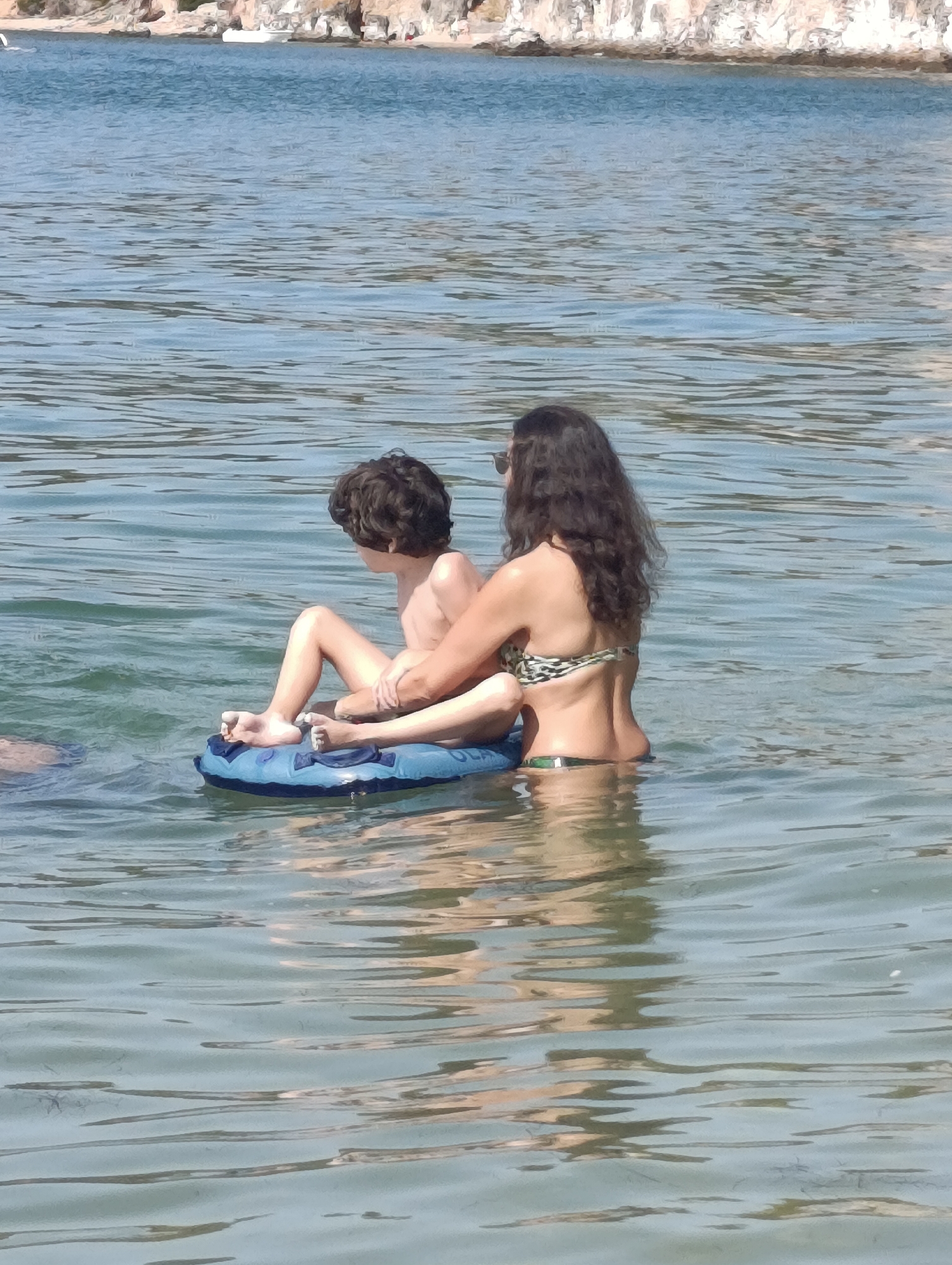 A woman and a young boy are in the water near a rocky shore. The woman is wearing a bikini and sunglasses. She is holding the boy, who is sitting on an inflatable ring. The water is clear and blue, and the sun is shining. The rocky shore in the background appears to be a beach with some vegetation. The scene suggests a relaxed day at the beach, possibly during the summer. The time of day appears to be midday, given the bright sunlight and the shadows. The overall mood is peaceful and tranquil. There is no visible text in the image.