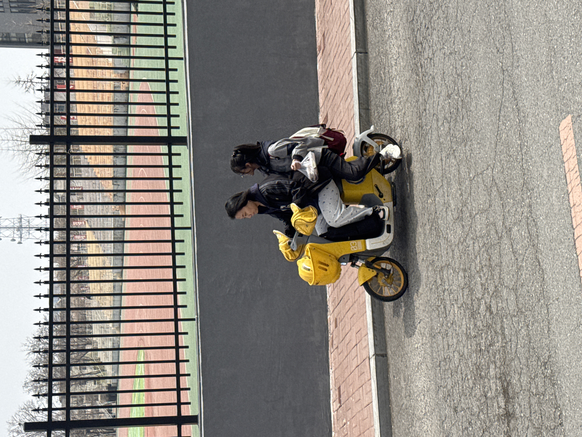 Two young women are seen riding a bright yellow electric scooter on a paved road in Zhongtun, China, during the daytime under an overcast sky. The woman in front wears a dark jacket, light-colored patterned pants, and a yellow fanny pack, with her dark hair pulled back. Behind her, the second woman is dressed in a gray and white jacket, dark pants, and glasses, carrying a maroon backpack and holding a white plastic bag. Both appear to be focused on riding.
To the left of the road, a black metal fence with pointed decorative tops acts as a barrier to a sports field or stadium. Through the fence, a red-brown track and green turf are visible. Beyond the field, leafless trees and some building structures or scaffolding can be discerned in the background, suggesting a cooler season. The road itself is asphalt with visible cracks, bordered by a brick-colored sidewalk or pathway to the right. The yellow electric scooter has white accents and yellow wheels. The number "03" is visible on the front of the scooter, and a logo resembling two vertical parallel lines (||) is on its side. The scene captures an ordinary urban activity.