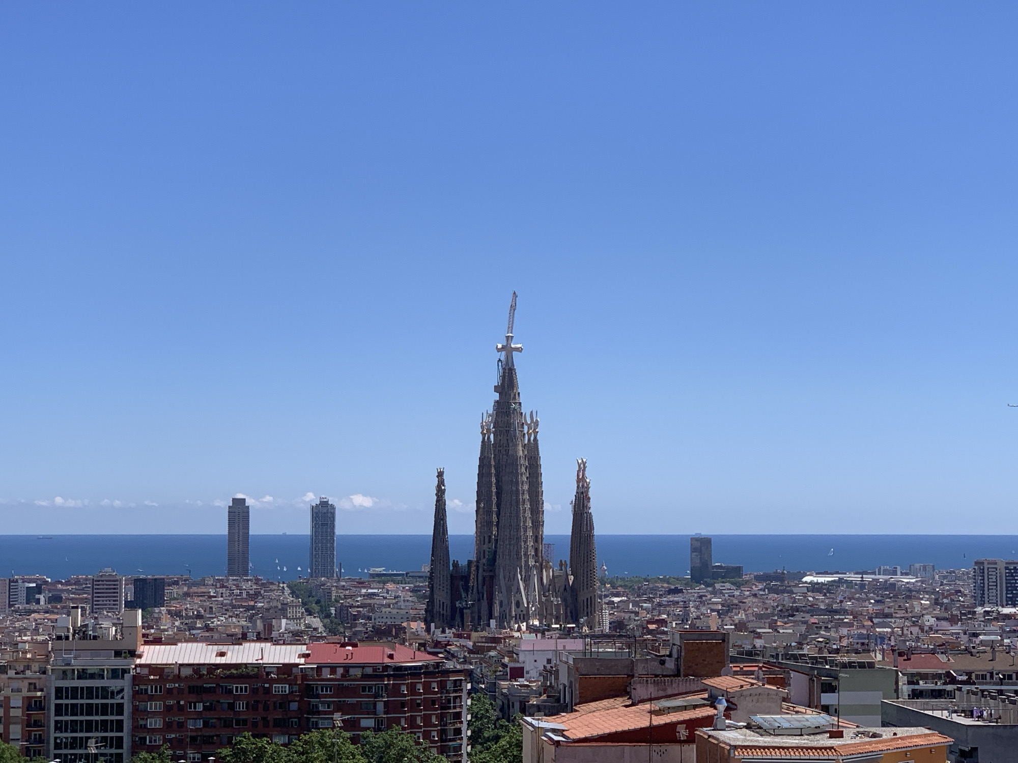 This image depicts an elevated view of Barcelona, Spain, showcasing the Sagrada Familia as the central architectural feature. The Sagrada Familia's iconic spires dominate the cityscape, with a construction crane visible at its peak. In the background, the Mediterranean Sea is visible on the horizon, dotted with distant ships. Several modern high-rise buildings are visible along the waterfront. The urban landscape consists of densely packed buildings with red-tiled roofs. The sky is clear and blue.