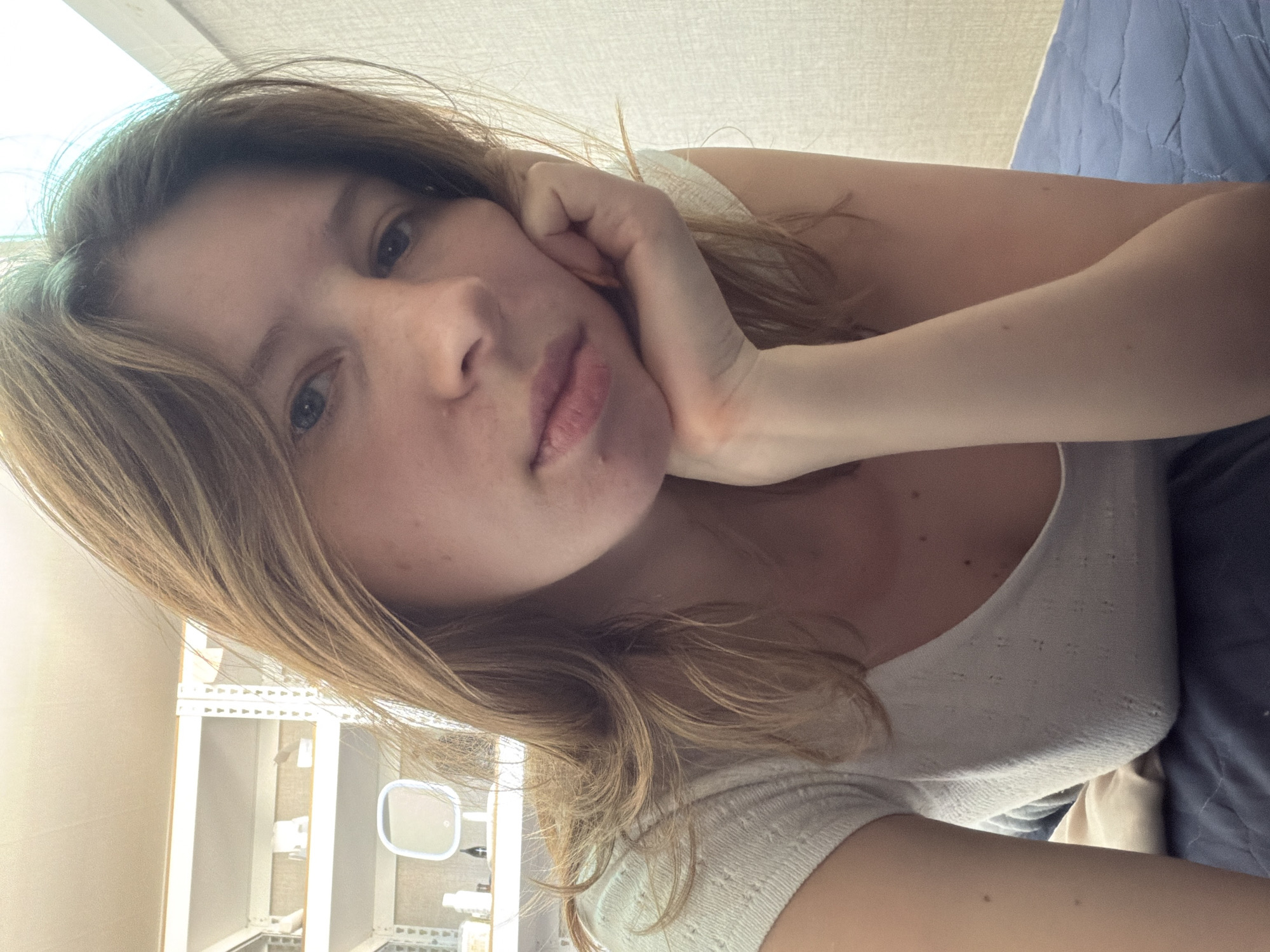 The image shows a young woman with light brown hair, resting her chin on her hand. She is wearing a light-colored, textured top. In the background, there are white shelves filled with various items, and a small, illuminated mirror. The setting appears to be an indoor space, possibly a bedroom or dressing area. The overall lighting suggests it could be daytime. There is no visible indication of the city of Dongducheon-si, South Korea.