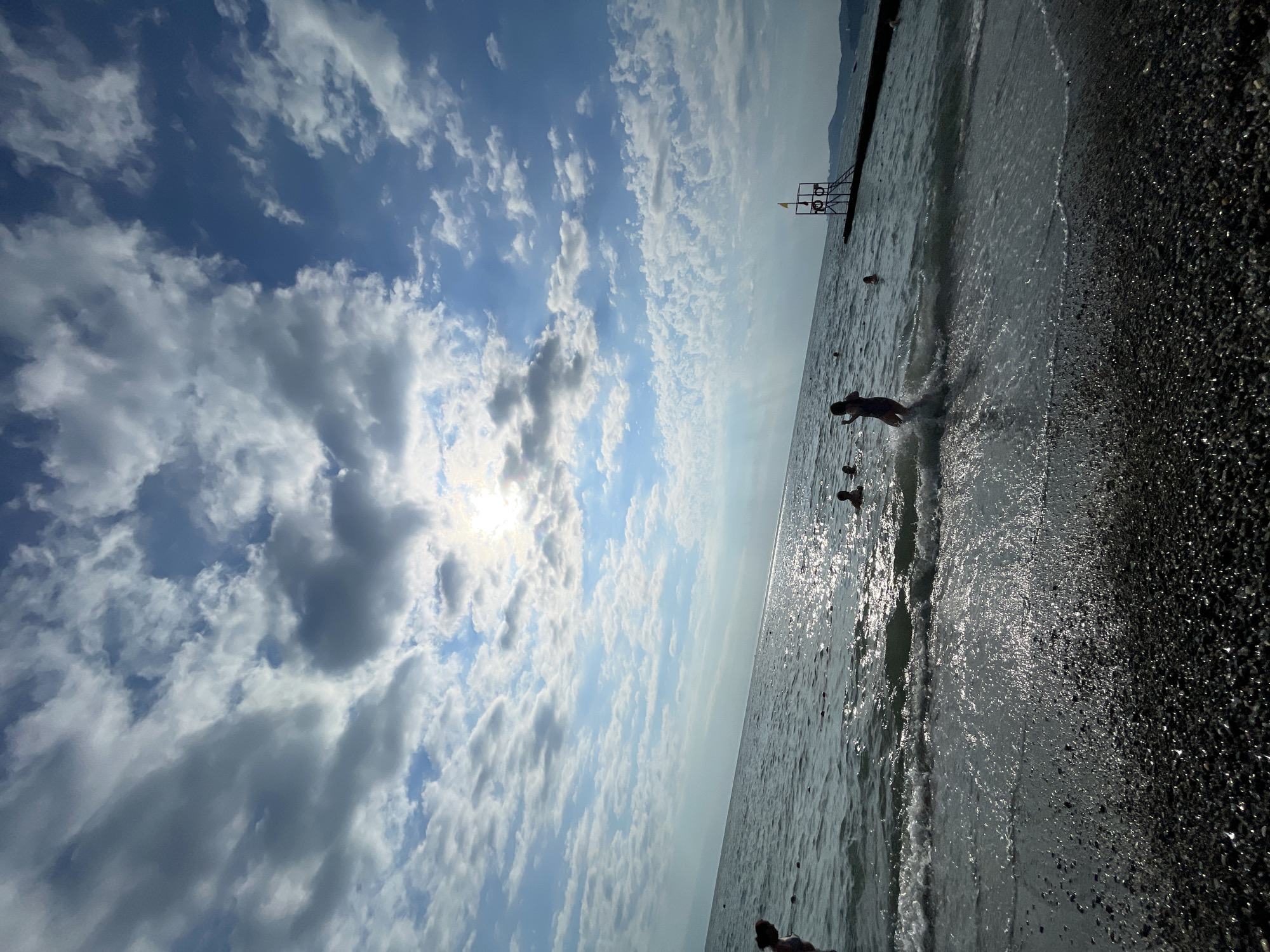 The image depicts a sunny day at a pebble beach in Sochi, Russia. The sky is filled with dramatic cumulus clouds, with the sun shining brightly through them. The sea is calm and glistening, with gentle waves lapping at the shore. Several people are visible in the water, enjoying a swim. In the distance, a lifeguard tower with a yellow flag stands, suggesting a designated swimming area. The pebbles on the beach are dark and reflective, catching the sunlight. The overall atmosphere is one of leisure and enjoyment on a beautiful summer day.