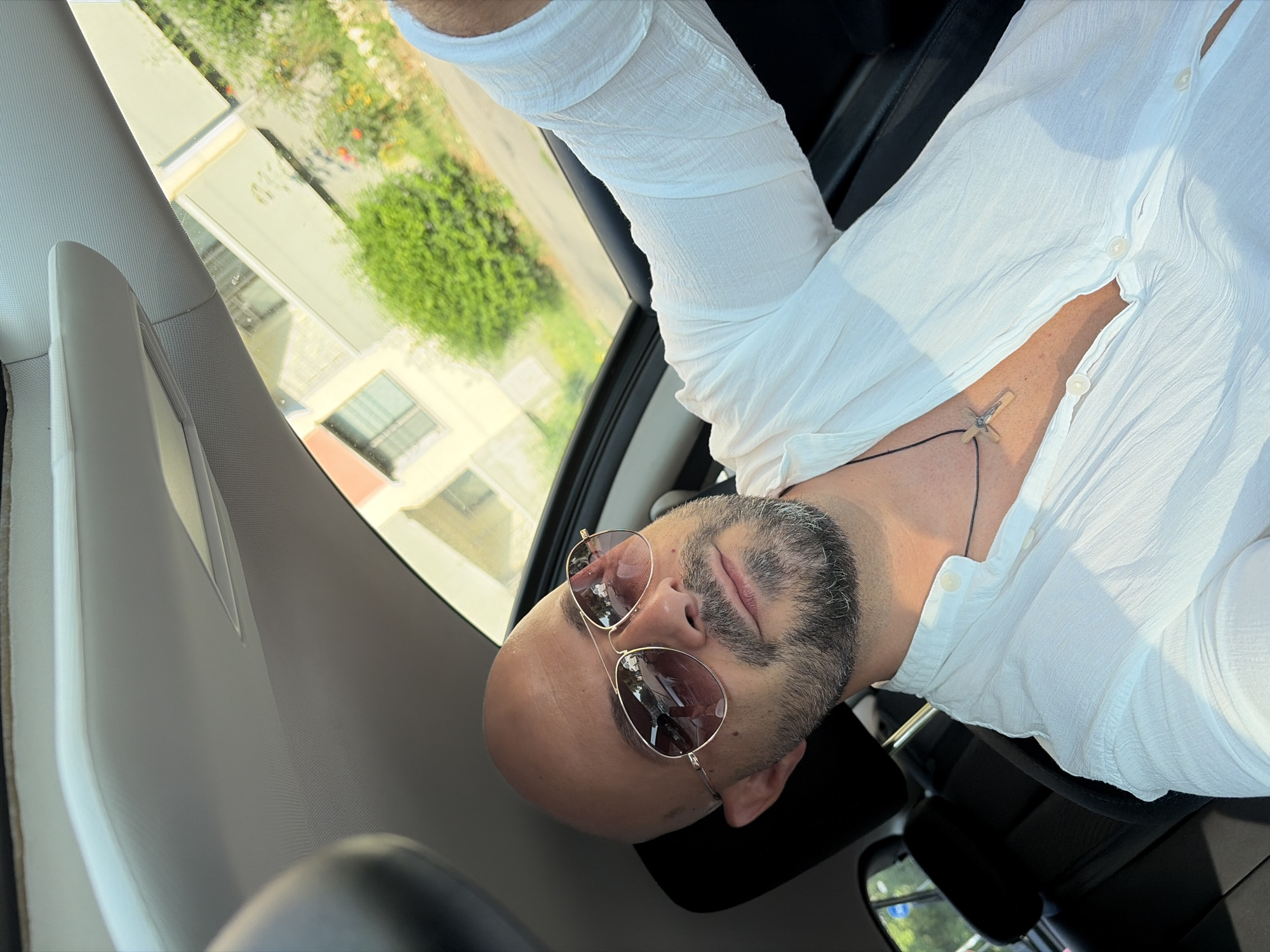A bald man with a goatee and sunglasses is in the driver's seat of a car, looking at the camera. He is wearing a white, open-collared shirt with a cross pendant. The car has a sunroof, and the exterior view shows a building and greenery. The image was taken in Sofia, Bulgaria.