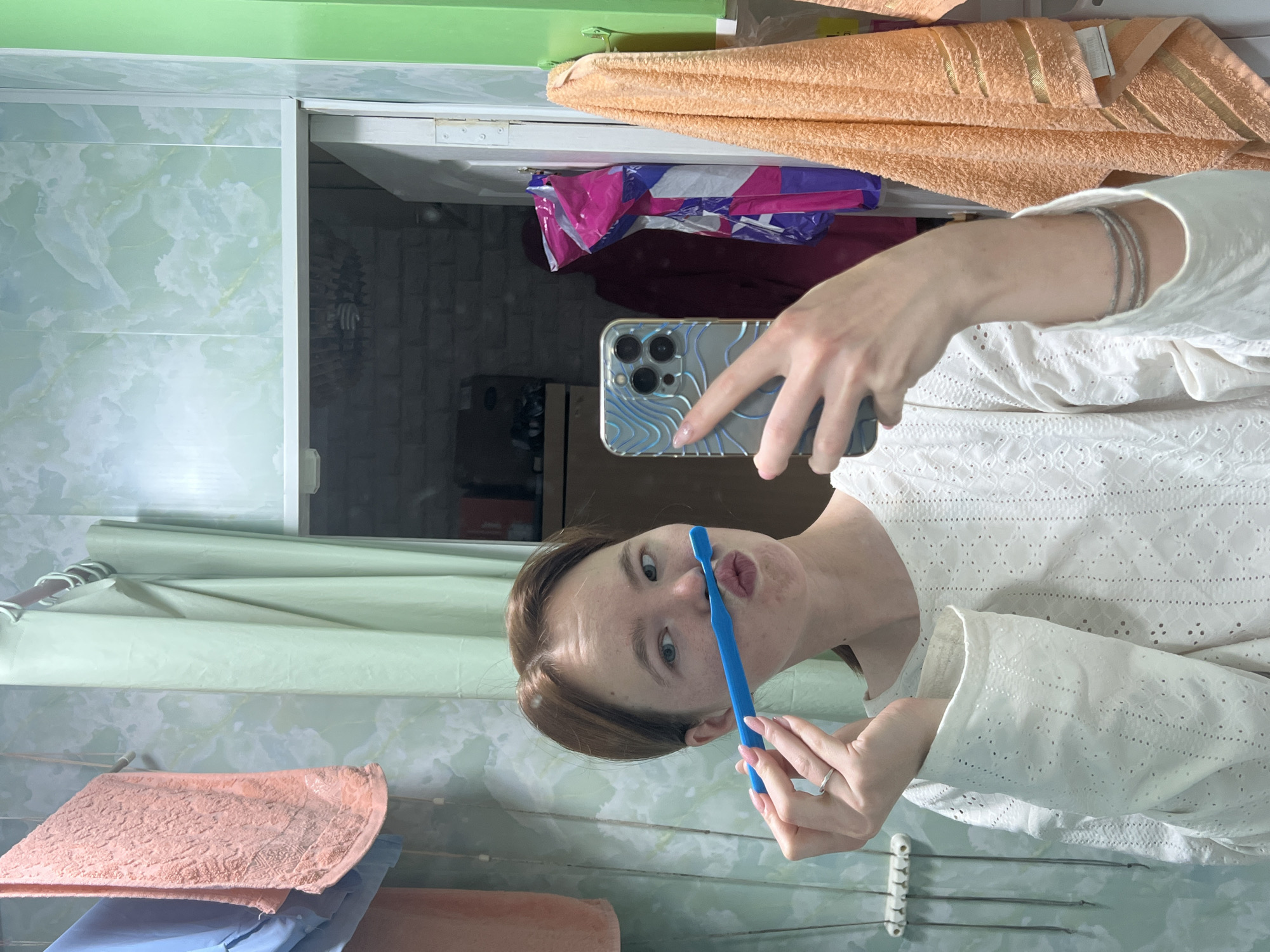 The image is a vertical mirror selfie taken by a young woman in a bathroom in Chaikovsky, Russia.
The central subject is a young woman with light brown hair and fair skin, wearing a white long-sleeved top with embroidered eyelet details. She is holding a blue toothbrush with both hands, pressing the bristles against her upper lip, and making a playful "fish face" or "duck face" with pursed lips and wide, light-colored eyes. Her left hand, wearing a silver ring, is near the toothbrush, while her right hand holds a silver iPhone (likely a Pro/Pro Max model) with a clear case featuring a light blue wavy, topographic pattern, used to capture the reflection. Several thin silver bracelets are visible on her left wrist.
The bathroom setting is reflected in the mirror. To the left, a light green wall panel or shower curtain is visible, contrasting with a brighter green painted ceiling or upper wall section at the top. An orange/peach colored towel hangs on a rack in the upper right, displaying a small, illegible white label. Another similar colored towel is folded in the lower left, partially obscuring a blue item. A white wall-mounted hanger rack with hooks is also seen in the lower left. Through an opening or at the edge of the mirror on the right, a darker room is reflected, revealing a wall with a brick-like texture and what appear to be brightly colored plastic bags or clothing (magenta, purple, blue) in the background. The overall mood conveyed by the woman's expression is playful and lighthearted.