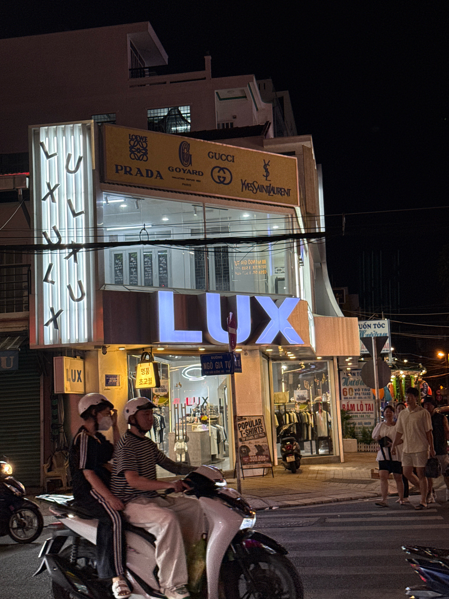 The image shows a street scene at night in Nha Trang, Vietnam, likely on a busy commercial street. In the foreground, two people are riding a scooter. The person in the front is wearing a white helmet and a black and white striped shirt, while the person in the back is wearing a white helmet, a black tracksuit, and white sandals. They are on a light-colored scooter.
Behind the scooter, a building with multiple floors is visible. The lower floors house shops, with prominent signage. The most visible sign on the upper part of the building displays logos of luxury brands such as LOEWE, PRADA, GOYARD, GUCCI, and YVES SAINT LAURENT on a yellow background. Below this, the word "LUX" is illuminated in large, blue, capital letters. The ground floor of the building appears to be a store, possibly selling clothing, with racks of garments visible through the windows. A sign for "POPULAR T-SHIRTS" with a price of "300.000" is displayed outside the store.
Further to the right, other shop signs are visible, including "VUON TOC" and "NHAT TRANG". Several people are walking on the sidewalk in the background, and streetlights illuminate the scene. The street is paved, and a crosswalk is present in the foreground. Various motorbikes are parked or passing by. The overall impression is a lively urban environment at night.