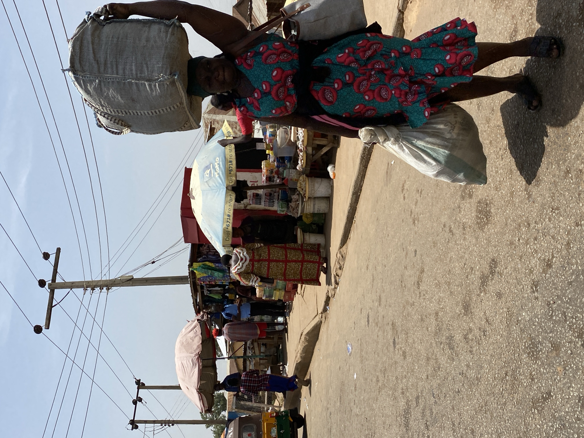 In Bukuru, Nigeria, an older woman is carrying a large sack on her head and another in her hand, walking on a paved road alongside a market. She is wearing a green and red floral dress and sandals. Several market stalls with umbrellas and awnings are visible, filled with people. Electrical poles and wires line the street. A three-wheeled vehicle can be seen in the lower left.