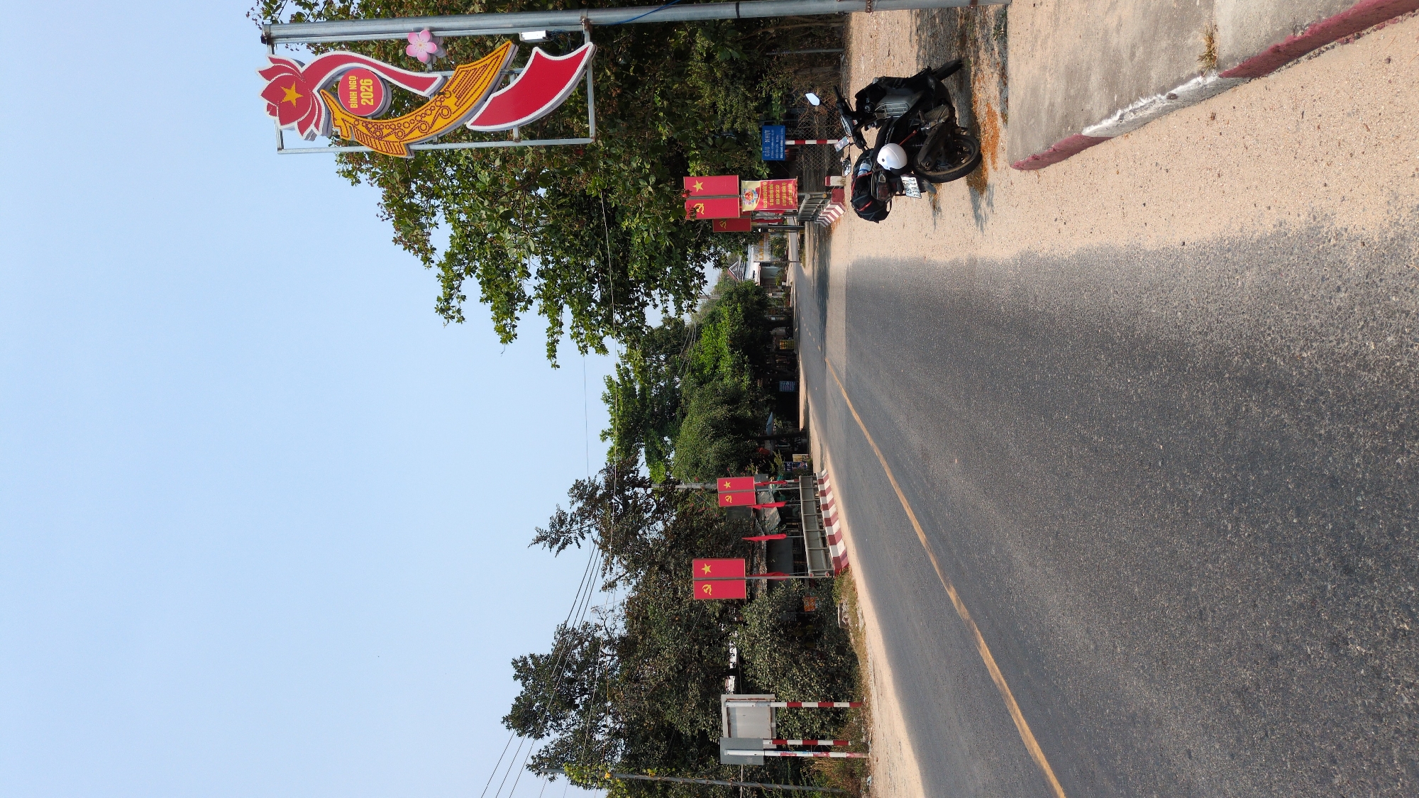 The scene depicts a street in Lâm Sơn, Vietnam. A black motorcycle is parked on the right side of the road, next to a sidewalk with a red and white curb. The road is paved with asphalt and has a yellow line marking the center. Along the left side of the road, there are several red flags with a hammer and sickle emblem. A large sign with Vietnamese script and decorative elements is also visible on the left. Trees line both sides of the street, and the sky is clear and blue.