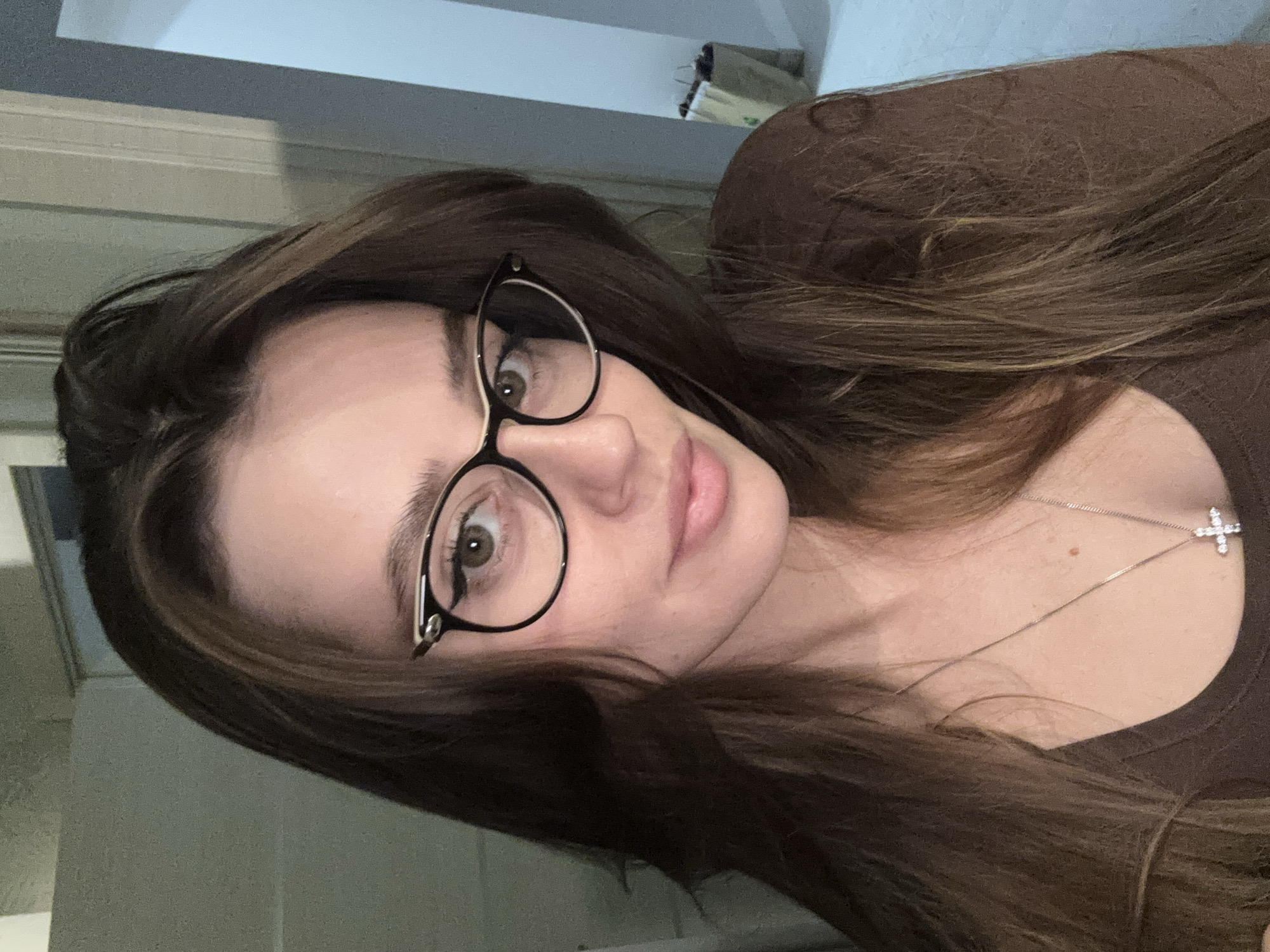A woman with long, brown hair is facing the camera, wearing glasses and a brown shirt. She also has a silver necklace with a cross pendant. The background includes a light-colored wall and a glimpse of a brown bag. It is not possible to identify a location in Krasnoyarsk, Russia from this image.