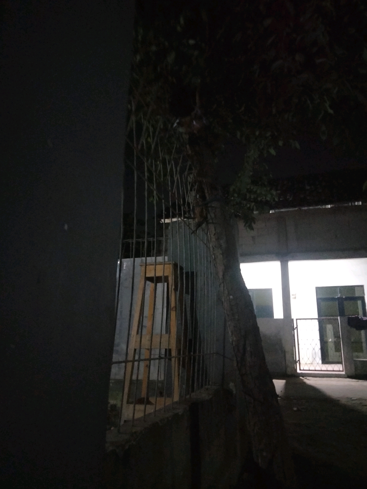 The image shows a dimly lit outdoor scene, likely at night, in what appears to be a residential or institutional area in Suka Sari, Indonesia. A large, dark wall dominates the left side of the frame. To the right of the wall is a metal fence. Behind the fence, a wooden structure, possibly a stool or a small table, is visible. A tree with a thick trunk and dark foliage grows alongside the fence, extending into the upper portion of the image. Further to the right, a building with illuminated windows and a decorative metal gate is visible. The ground is dark and appears to be paved or covered with dirt. Shadows are cast across the ground, indicating a light source from the building. No people are clearly visible in the image.