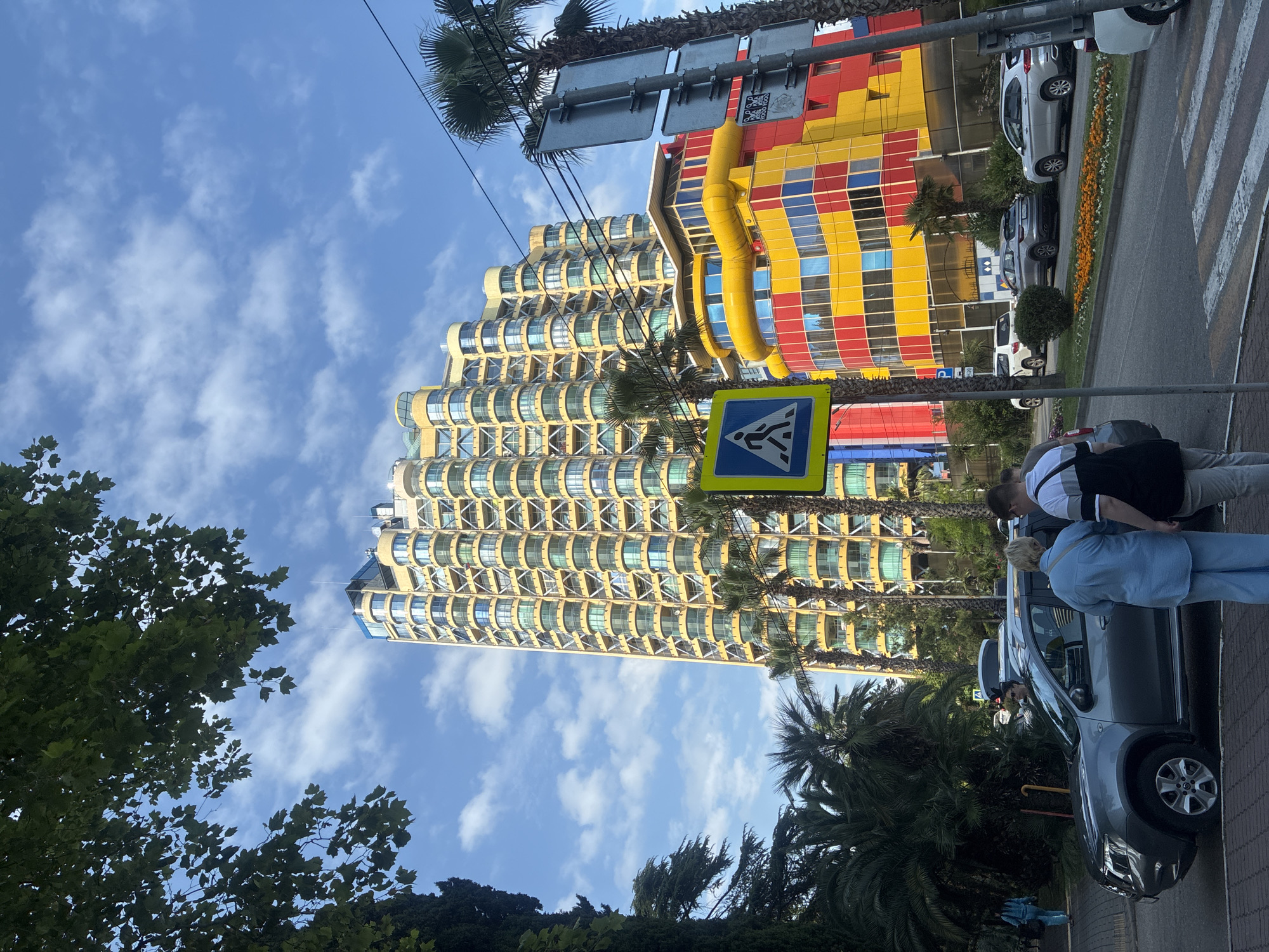 The image depicts a street scene in Sochi, Russia, with a focus on two distinct architectural styles. On the right, a building with a vibrant, checkerboard facade of red, yellow, and blue panels stands out. To its left, a towering, curved building with a distinctive yellow, layered design features numerous balconies with glass railings, creating a wave-like appearance.
In the foreground, several people are visible near a gray car parked on the side of the road. A pedestrian crossing with white stripes is also present. A blue pedestrian crossing sign with a white pictogram of a person walking is prominently displayed. Palm trees and other lush green foliage are scattered throughout the scene, characteristic of a warm climate. The sky is blue with scattered white clouds, suggesting it is daytime. The overall atmosphere appears casual and urban, with elements of modern and perhaps resort-style architecture. There is no visible text in the image.