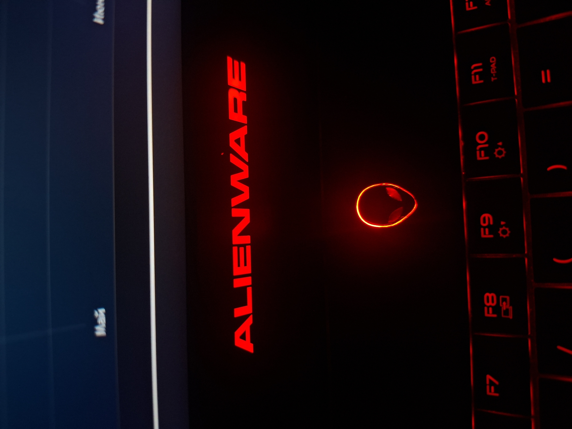 The image shows a close-up of an Alienware laptop with red illuminated accents. The Alienware logo is prominently displayed in red on the lid. Below the logo, the Alienware's iconic alien head symbol is also illuminated in red, situated above the keyboard. The keyboard itself is black with red backlighting, and several function keys (F7, F8, F9, F10, F11) are visible. The overall setting appears to be indoors, with dim lighting contributing to the glowing effect of the laptop's features. There is no visible indication of the city of Užuežerė, Lithuania, within the image.