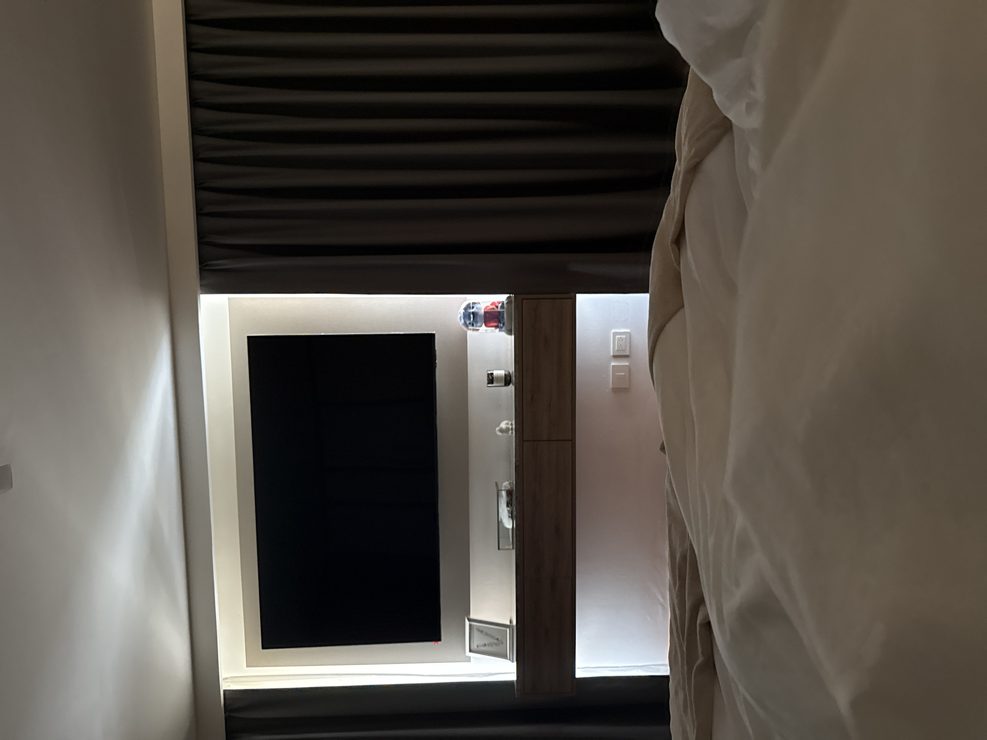 This image captures an interior scene, likely a modern hotel room or living space, viewed from a bed. The original image is rotated 90 degrees counter-clockwise.
Upon re-orientation, the upper portion of the frame features dark, horizontal blinds, largely drawn to cover a window. Below these blinds, a large, wall-mounted flat-screen television with a black screen, indicating it is off, is centrally positioned. To the right of the television, a tall, narrow wooden cabinet with internal shelving is visible. Several items are displayed within the cabinet, including what appears to be a wine bottle and other containers. Above the cabinet, two plastic bottles, one red and one blue, are also visible.
The area around the television and cabinet is subtly illuminated by strip lighting or backlighting, creating a contemporary ambiance. On the light-colored wall to the far right, a series of white light switches or power outlets are installed. The right side of the frame is dominated by soft, light-colored bedding, likely a duvet or sheets, suggesting the photo was taken from a bed within the room. The walls are predominantly light, possibly white or light gray, with gentle reflections from the lighting on the left wall.
The scene is devoid of people and there is no discernible activity. The lighting suggests it could be nighttime or that the blinds are drawn to dim the room. The overall aesthetic is clean and modern. No visible text is present. This interior setting is located in San Bernardino Tlaxcalancingo, Mexico.