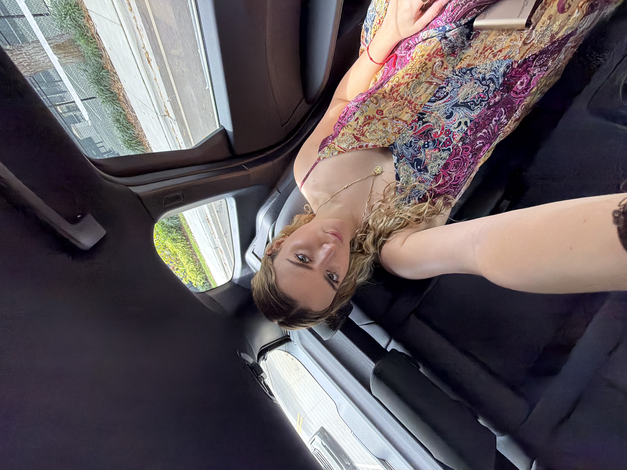 A young Caucasian woman with blonde hair is looking into the camera inside a car, possibly a rideshare, in Key Biscayne, United States of America. She is wearing a colorful floral-patterned dress and a delicate gold necklace. She is sitting in the back seat, as evident from the car's interior features, including the black upholstery and door trim. Through the car windows, vegetation and a paved road are visible. She appears to be taking a selfie with her arm extended toward the camera.