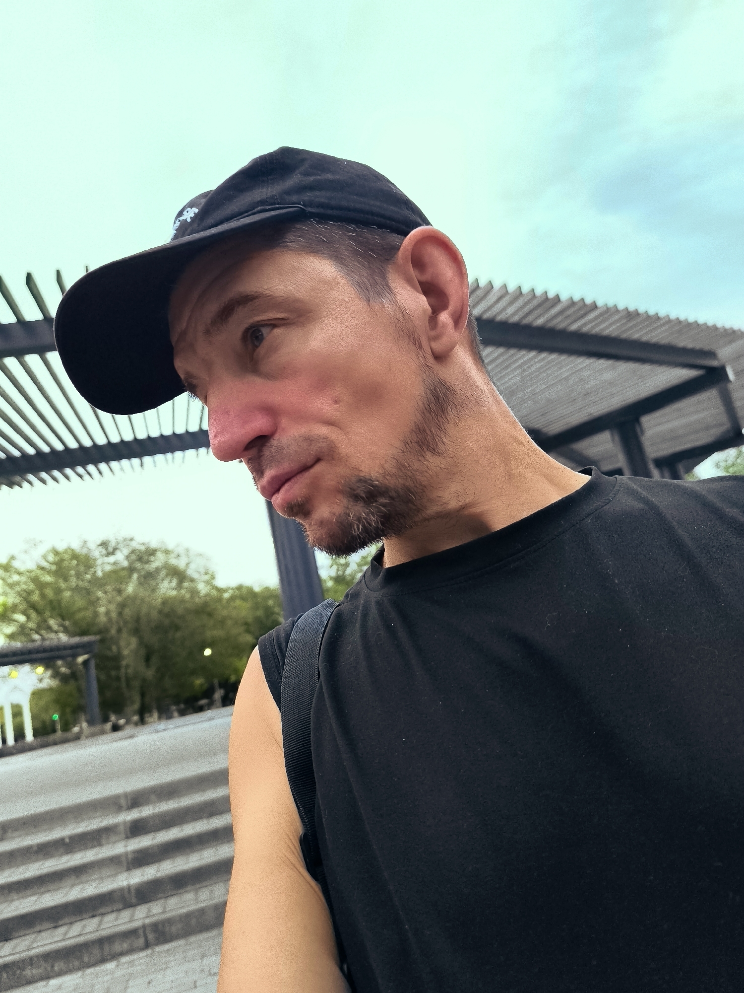 A man wearing a black baseball cap and black t-shirt is pictured from the chest up, looking downwards and to his left. He has short brown hair and stubble on his chin and jawline. His left ear is visible. In the background, a grey, corrugated awning or roof structure is visible. Below that, trees and a white building with arches can be seen. To the left of the man, a set of grey concrete stairs descends. The sky is a pale blue with some clouds. The man appears to be outdoors in a park-like or public space. The location is identified as the city administration of Qarağandı city, Kazakhstan.