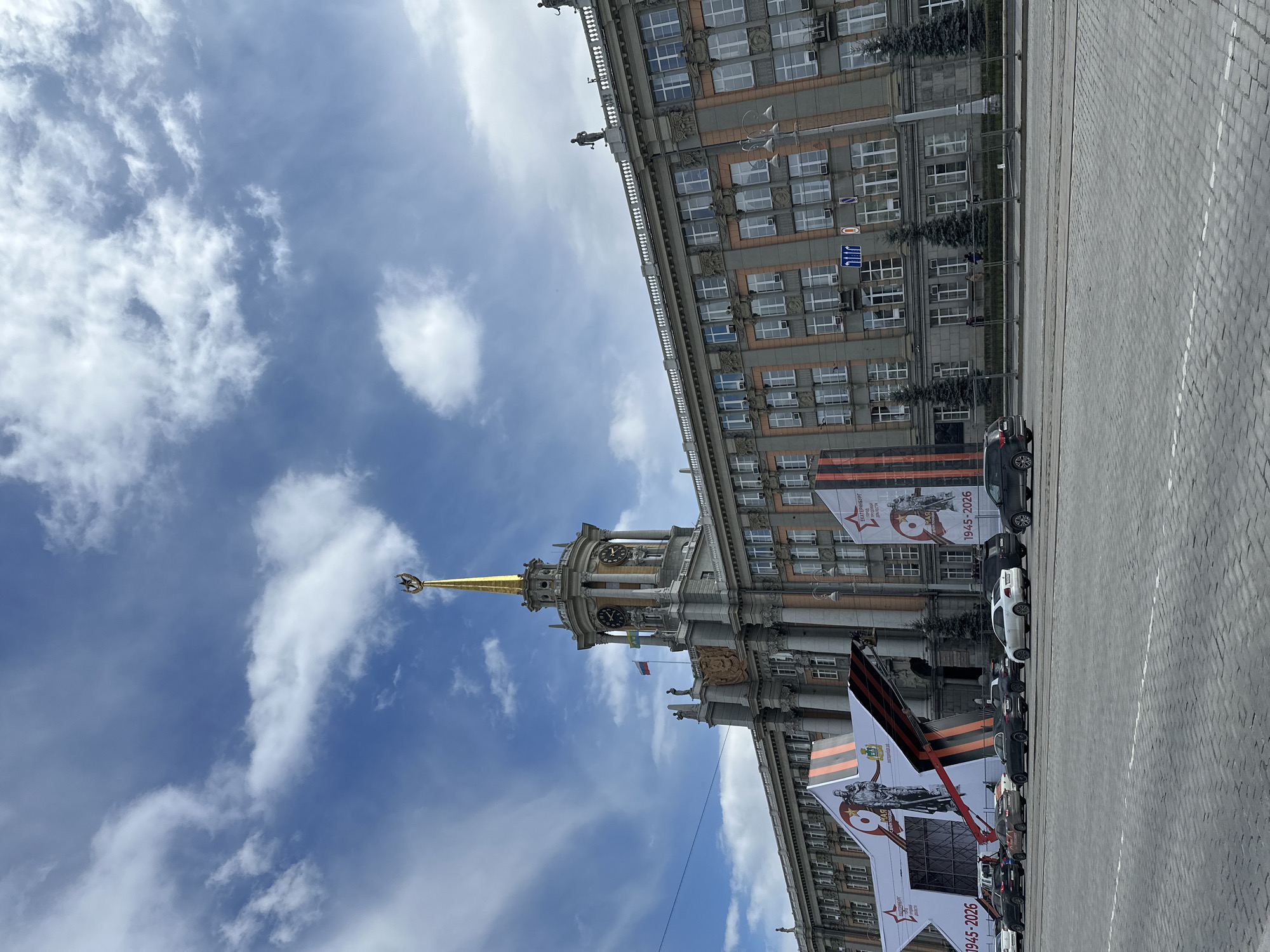 The image shows the City Hall building in Yekaterinburg, Russia, taken from a ground-level perspective looking upwards. The building has a tall spire topped with a golden Soviet star and is adorned with banners commemorating Victory Day, marked with "1945-2026." Several vehicles are parked along the cobblestone street in front of the building. The sky above is partly cloudy.