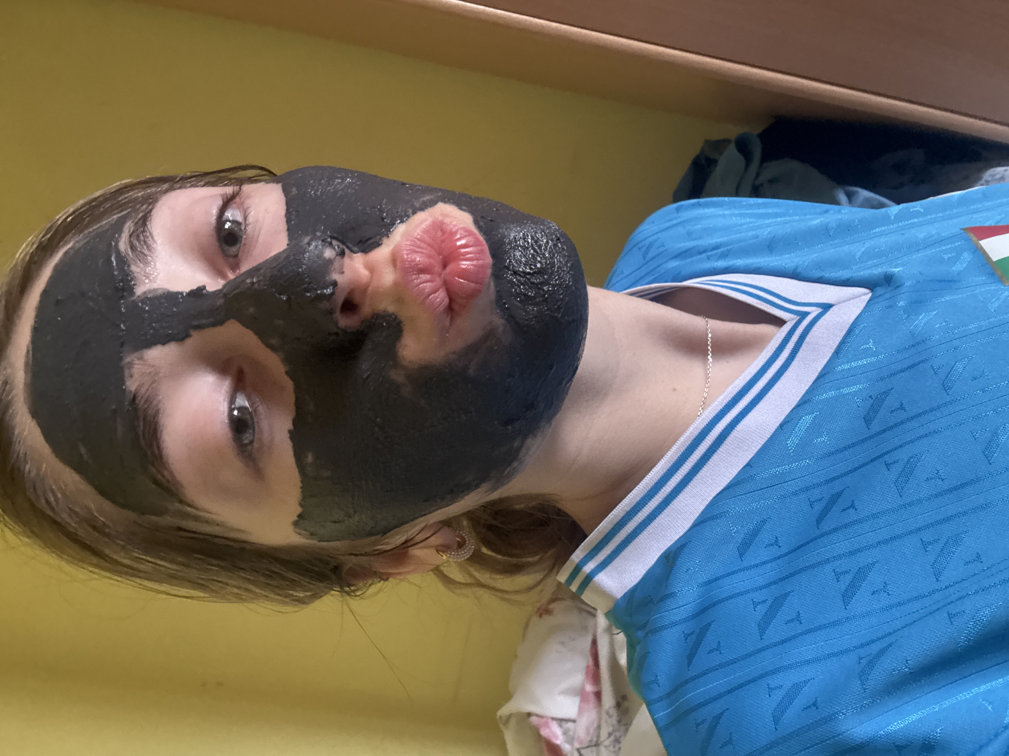 A woman is wearing a blue jersey with white trim around the neck and sleeves, and a Hungarian flag patch is visible on the right side of the jersey. Her face is covered in a dark substance, possibly a facial mask, except for the lips. She has light skin, light brown hair, and is wearing hoop earrings. A gold necklace is visible on her neck. The background includes a yellow wall and a wooden shelf or ledge. Other objects are visible. The location is Unknown, Unknown.