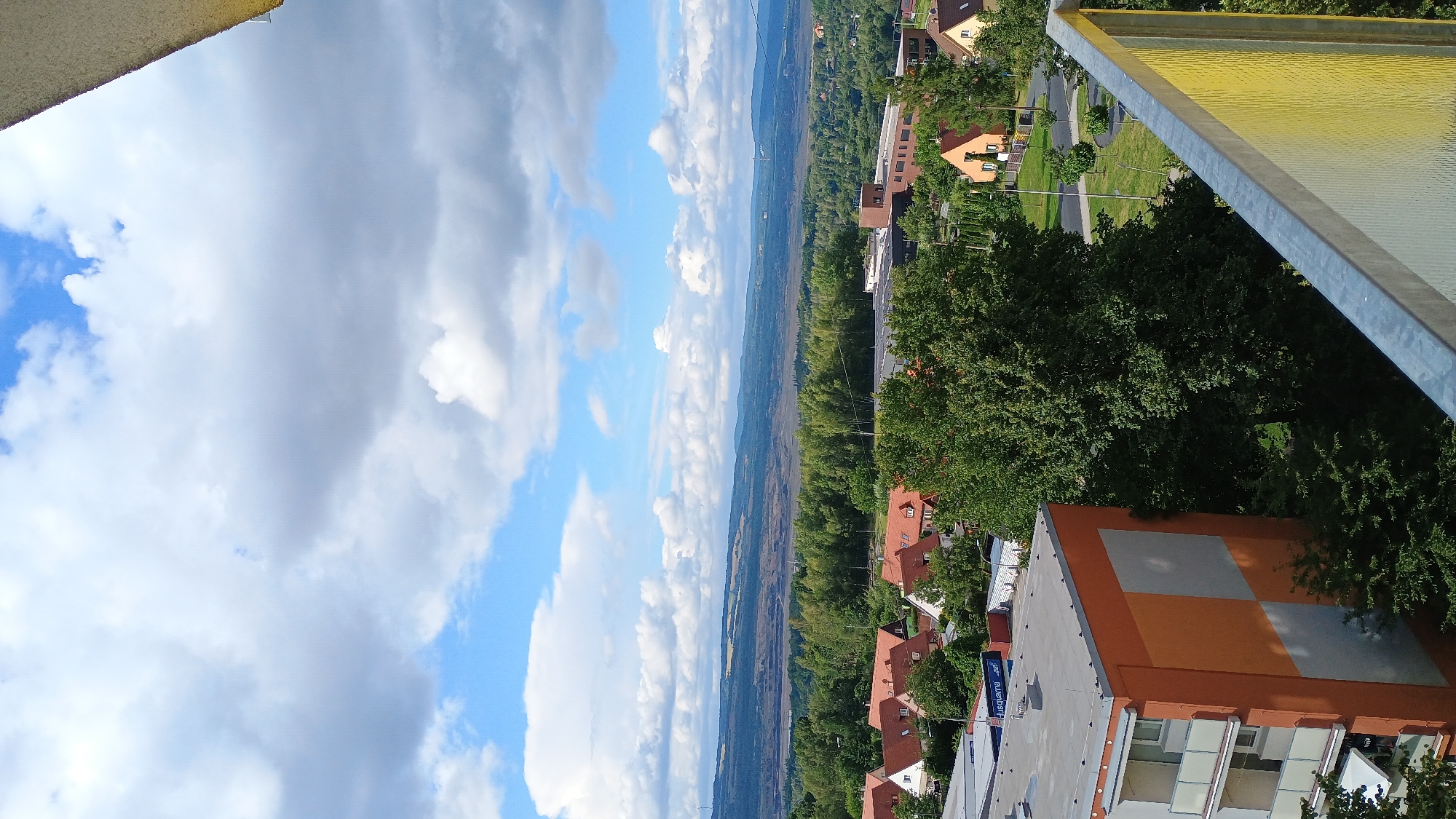 The image is an elevated view of the city of Sokolov, Czechia, on a partly cloudy day. In the foreground, the corner of a building with an orange and gray facade is visible, along with a metal railing. Behind this building, a dense cluster of green trees dominates the mid-ground, obscuring some of the residential area below. The residential area consists of several houses with red tile roofs, interspersed with more greenery and paved roads. Further in the distance, a large, open-pit mine, characteristic of the Sokolov region's industrial past and present, is visible. Beyond the mine, rolling hills and mountains stretch across the horizon under a sky filled with scattered white and gray clouds against a bright blue backdrop. No people are visible in the image.