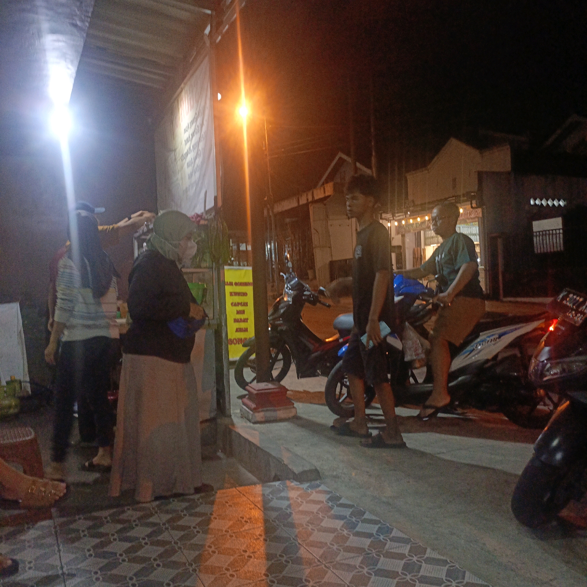 The image depicts an outdoor scene at night, likely in an urban environment. Several individuals are present, along with motorcycles and buildings in the background. The lighting suggests a street or market setting, possibly illuminated by streetlights and the lights from nearby establishments. The presence of a yellow banner with text in Indonesian, along with the general atmosphere, indicates the location is likely in Salatiga, Indonesia.
In the foreground, a woman wearing a headscarf and a mask is visible, standing next to a display or counter. To her left, another person is partially visible, wearing a striped shirt and dark pants. Further to the right, a young man in a black t-shirt is standing, and another man, wearing glasses and a green t-shirt, is seated on a motorcycle. A motorcycle is also parked in the immediate foreground, partially obscuring the lower portion of the scene. The ground is tiled with a geometric pattern.
In the background, buildings with windows and signs are visible. A large white banner with text is hung on a structure, and a bright light source is illuminating the area from the left. Another yellow banner with text is situated on a pole near the center of the image. The overall impression is a bustling nighttime scene, possibly a food stall or small market. The presence of Indonesian language on signage confirms the location as Indonesia, and the specific context points to Salatiga.