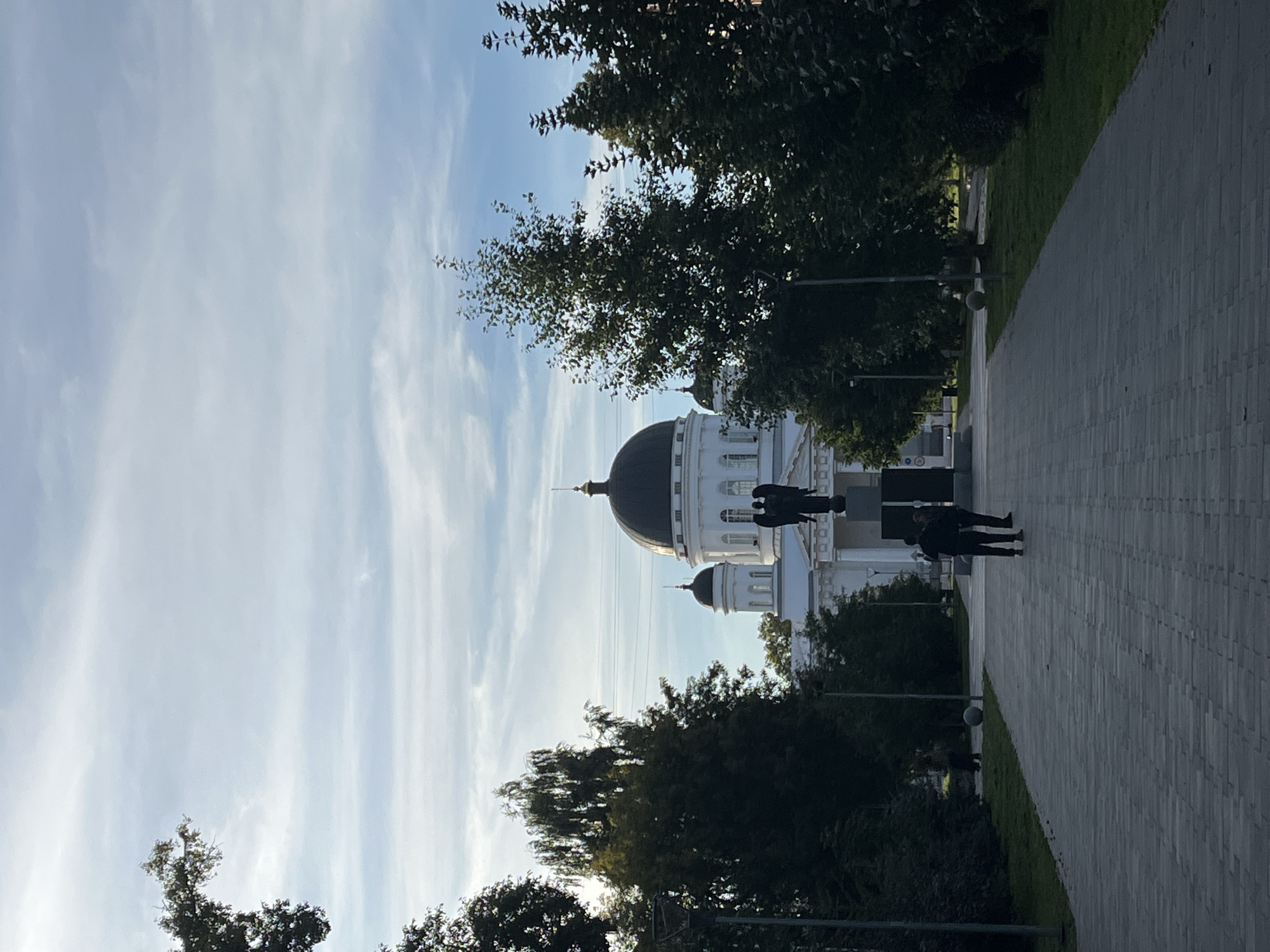 The image depicts a view of the Alexander Nevsky Cathedral in Nizhny Novgorod, Russia, on a cloudy day. In the foreground, a wide paved walkway leads towards the cathedral. Two figures, silhouetted against the bright sky, are walking away from the camera on the walkway. To the right of the walkway, a lush green lawn is visible, bordered by trees. A lamppost stands on the right side of the walkway. Further down the walkway, a statue of an angel is prominently displayed in front of the cathedral. The cathedral itself is a large, white building with a dark dome and multiple smaller domes. The sky is partially obscured by light, wispy clouds.