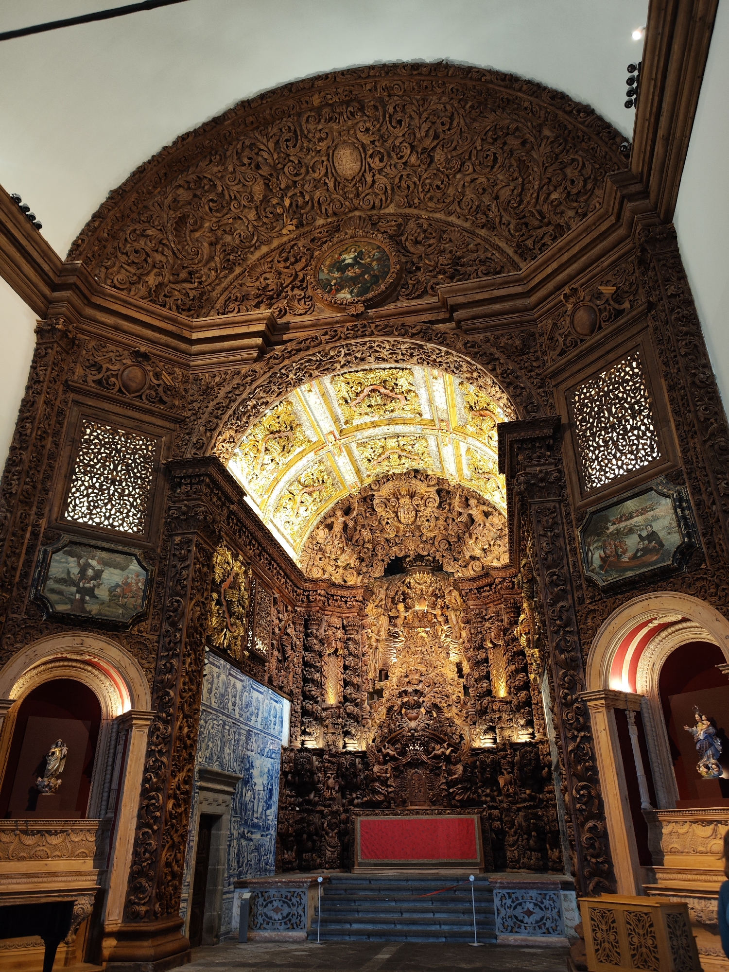 This image captures the richly decorated interior of a Baroque-style church or chapel, likely in Ponta Delgada, Portugal, given the architectural elements. The central focus is the altar, dominated by an incredibly ornate and gilded wooden altarpiece (retable) that reaches towards the high, vaulted ceiling. This altarpiece is characterized by intricate carvings, figures, and architectural motifs, with much of it highlighted in gold leaf, giving it a luminous appearance.
The walls and structures surrounding the altar are also extensively covered in carved dark wood, featuring elaborate floral patterns, volutes, and various decorative elements. Above the main arch of the altarpiece, a large, semicircular wooden panel displays more intricate carving, and a small circular painting is embedded within it at the apex. On either side of the main altar, there are large rectangular openings covered by finely carved wooden grilles, allowing light to filter through or serving as decorative elements. Below these grilles, two paintings depicting religious scenes are set into the wooden panels.
To the left of the altar, a large section of the wall is covered with traditional Portuguese blue and white azulejo tiles, depicting narrative scenes. Further out on both sides, arched niches house statues; the one on the left clearly shows a statue of the Virgin Mary holding the infant Jesus. The altar area itself features a red fabric panel and is approached by several stone steps. The ceiling above the main altar is also vaulted and richly decorated with gilded and painted designs. Artificial lights illuminate the intricate details of the woodwork and gilding.
Visible text in a cartouche within the upper carved wood panel appears to include Latin words, with "LVS" and "DEI" somewhat discernible.