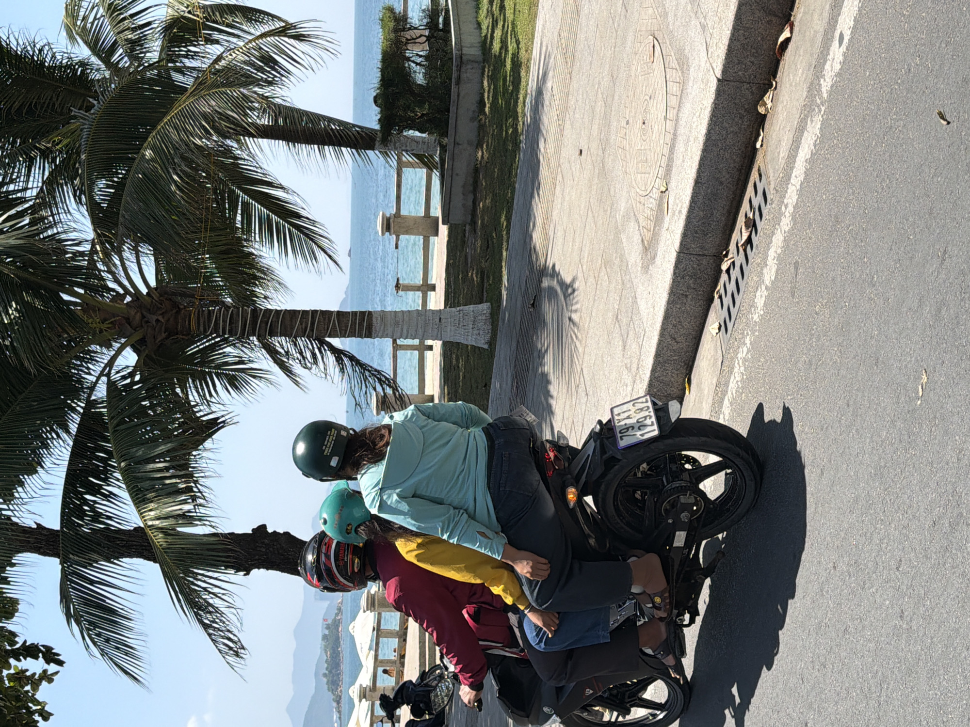 In Nha Trang, Vietnam, three individuals are riding a motorcycle on a paved road beside a sidewalk and a grassy area. The motorcycle has a license plate that reads "79-X1 129.82". The rider in the back is wearing a red and black helmet, the middle passenger is wearing a teal helmet, and the person in front is wearing a dark green helmet. The middle passenger is wearing a yellow jacket, and the person in front is wearing a light blue long-sleeved shirt and blue jeans. Palm trees are visible in the background, along with the ocean and a distant island. The day is sunny with a clear blue sky.