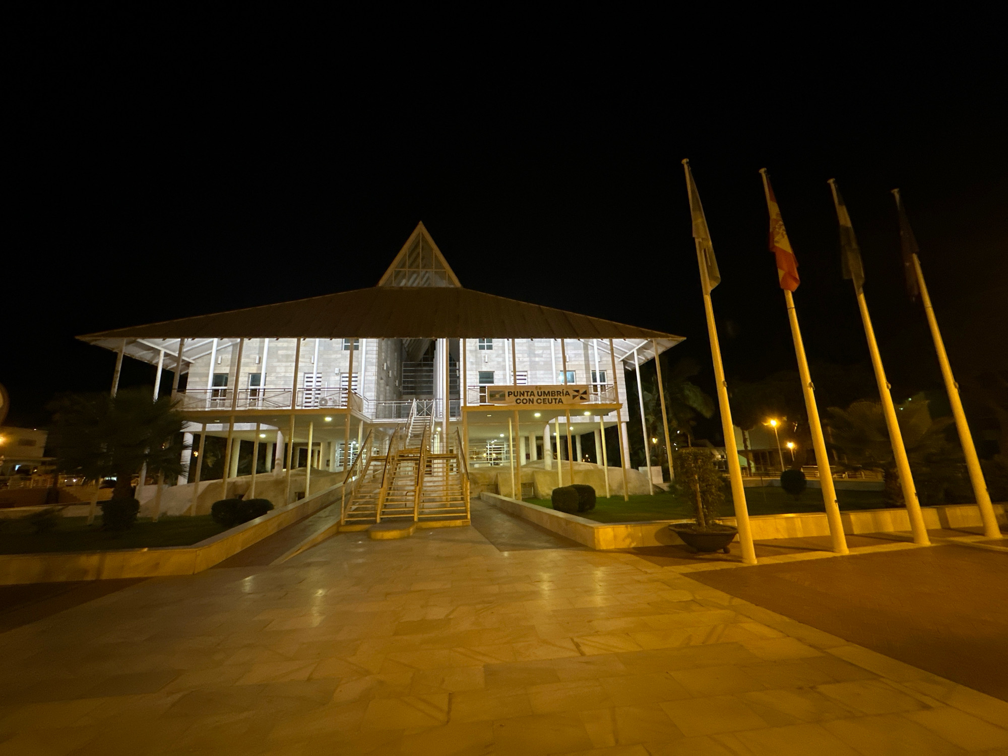 The image shows a building at night in Punta Umbría, Spain. The building is a multi-story structure with a distinctive pyramidal roof and white pillars supporting cantilevered floors. There are stairs and a ramp leading up to the building. Four flagpoles with flags are visible to the right of the building. The area in front of the building is paved with light-colored tiles and there are some trees and green spaces. The sky is dark, indicating nighttime. A sign on the building reads "PUNTA UMBRÍA CON CEUTA".