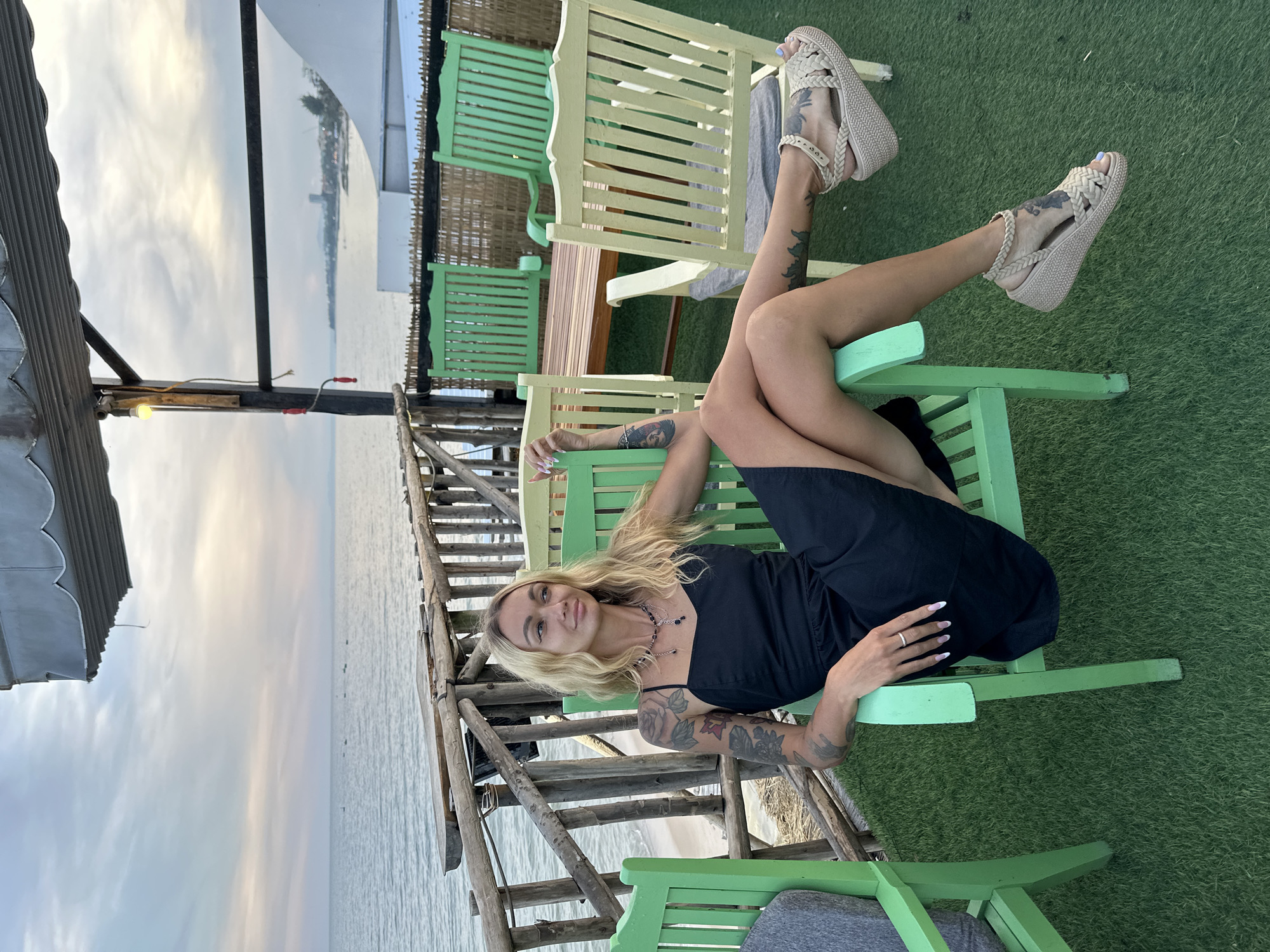 A woman with blonde hair and tattoos on her arms and legs sits in a green chair on a deck overlooking the ocean. She is wearing a black dress and tan sandals. The deck has a wooden railing and several other chairs, some green and some yellow. In the background, there is a wooden table and chairs, and beyond that, the ocean and a hazy shoreline with buildings. The sky is cloudy, with a mix of grey and pale pink clouds. The location is identified as Phu Thuy Ward, Vietnam.