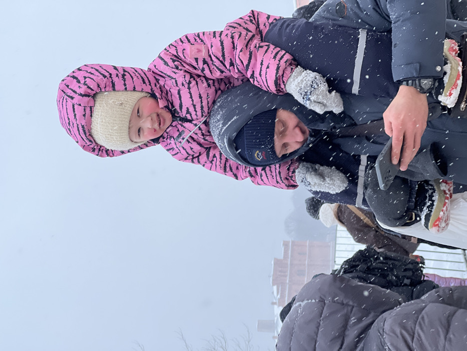 A person carries a child piggyback style in a snowy outdoor setting. The child wears a pink and black striped snowsuit with a cream-colored knit hat. The adult wears a dark blue hooded coat and hat. Snowflakes are visible throughout the scene. A building and other people are partially visible in the background.
