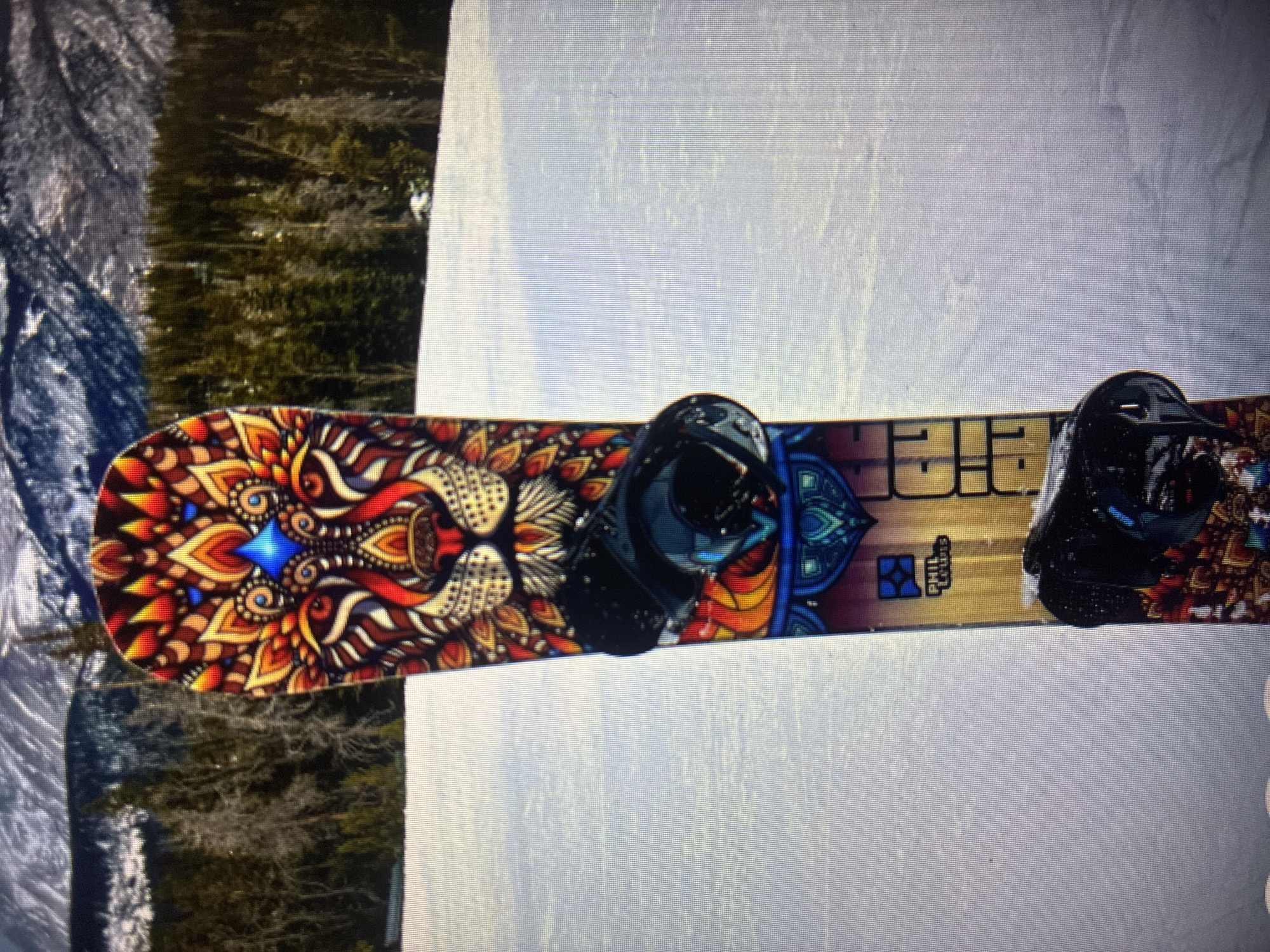 The image presents a colorful snowboard with two black bindings, resting on a snow-covered surface. The snowboard's top sheet features an elaborate and vibrant design, predominantly at the nose, depicting a stylized, mandala-like head, possibly a lion or mythical beast, rendered in rich tones of red, orange, yellow, blue, and black. Further down the board, toward the tail, additional intricate patterns and visible text "PHILLY" (or "PHILE") are discernible in a blocky, stylized font, accompanied by a smaller emblem.
The setting appears to be a winter landscape, likely a mountain slope or forest edge. To the left of the snowboard, a dense dark green treeline or forest is visible, while the rest of the background is dominated by white snow, indicating a clear, daytime scene. No individuals are present, and the snowboard lies stationary, suggesting it is either prepared for use, paused during an activity, or displayed. The overall image quality indicates it is a photograph taken of a digital screen, with a noticeable pixel grid pattern. This scene is consistent with a mountainous area near Asenovgrad, Bulgaria, known for its winter sports opportunities.