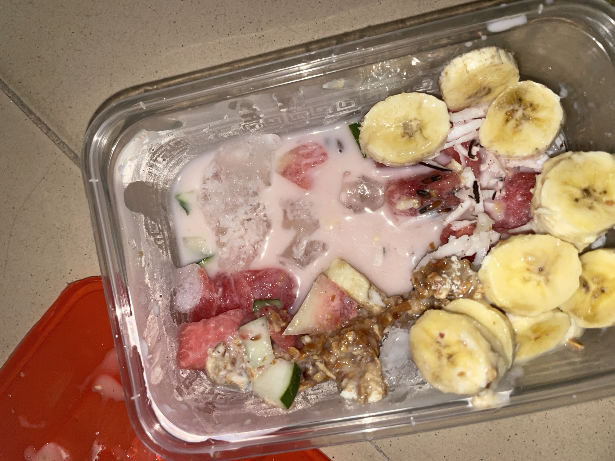 The image displays a clear plastic container viewed from above, holding a mixed food item. The container is partially filled with a pinkish-white liquid and contains several ice chunks. Slices of yellow banana, some exhibiting minor browning, are visible at the bottom and right side. Red fruit pieces, consistent with watermelon, are distributed in the upper and central portions of the mixture, some containing small dark seeds. Diced green pieces, resembling cucumber, are combined with a brownish, textured, granular substance on the left. White shredded material, indicative of coconut, is centrally placed, interspersed with a few dark, thin strands. A red plastic object, possibly a lid or another container, is partially visible in the upper-left corner of the frame. The container rests on a light-colored surface, likely a tiled countertop or table, with a grout line discernible in the upper right background. The scene is illuminated by artificial light. The image contains no visual elements that allow for the identification of a specific geographical location, such as the city of Jalingo, Nigeria.
