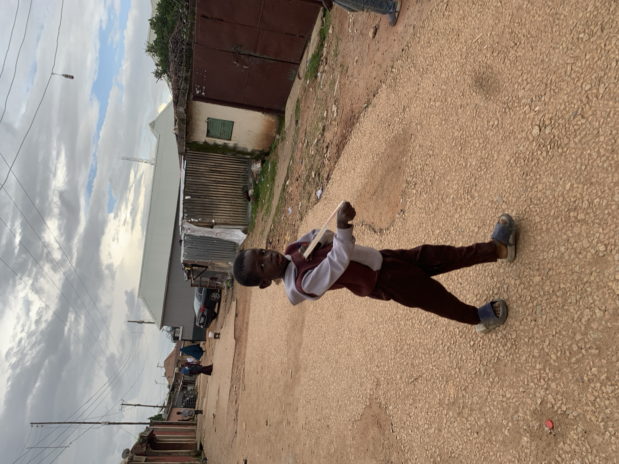In an outdoor scene on a cloudy day in the city of Jos, Nigeria, a young boy walks across a dirt road. He is wearing a maroon vest over a light-colored hoodie and dark pants, along with blue sandals. The boy is holding a piece of wood horizontally in front of him. The road is made of gravel and dirt, with some debris scattered on it. In the background, there are several buildings, including structures with corrugated metal roofs and walls, a dark-colored car, and a large brown gate. Overhead, a network of power lines is visible against the sky. Two other individuals are partially visible further down the road, one wearing a blue headscarf and the other in jeans.