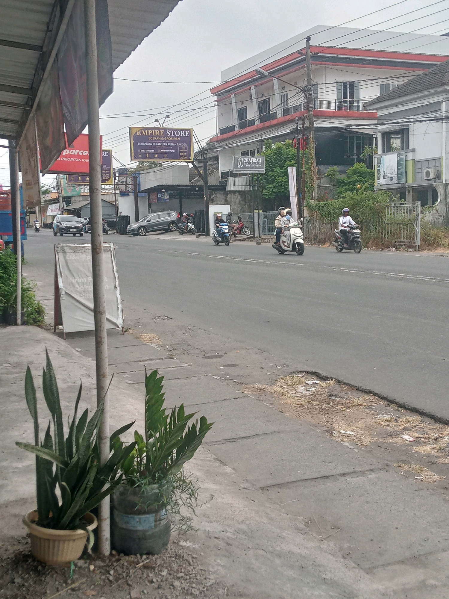 The image captures a street scene in Maguwoharjo, Indonesia, under an overcast sky. The foreground features two potted plants, one in a tan pot and another in a large recycled plastic bottle, situated on a concrete sidewalk. A metal pole stands between the plants.
The sidewalk leads to a two-lane asphalt road where several motorcycles are in motion. On the left side of the road, a black car is driving, followed by a gray car. Further down the road, two motorcycles are visible traveling in the same direction.
On the right side of the road, a white building with red trim dominates the background, featuring multiple floors and balconies. Several signs are visible on the building, including one for "EMPIRE STORE" that advertises "ECERAN & GROSIRAN SEMBAKO & PERABOTAN RUMAH TANGGA." Another sign displays the name "ZHAFIRAH."
A pedestrian crossing and a white fence are also present on the right side of the road. The overall environment suggests a commercial area with moderate traffic.