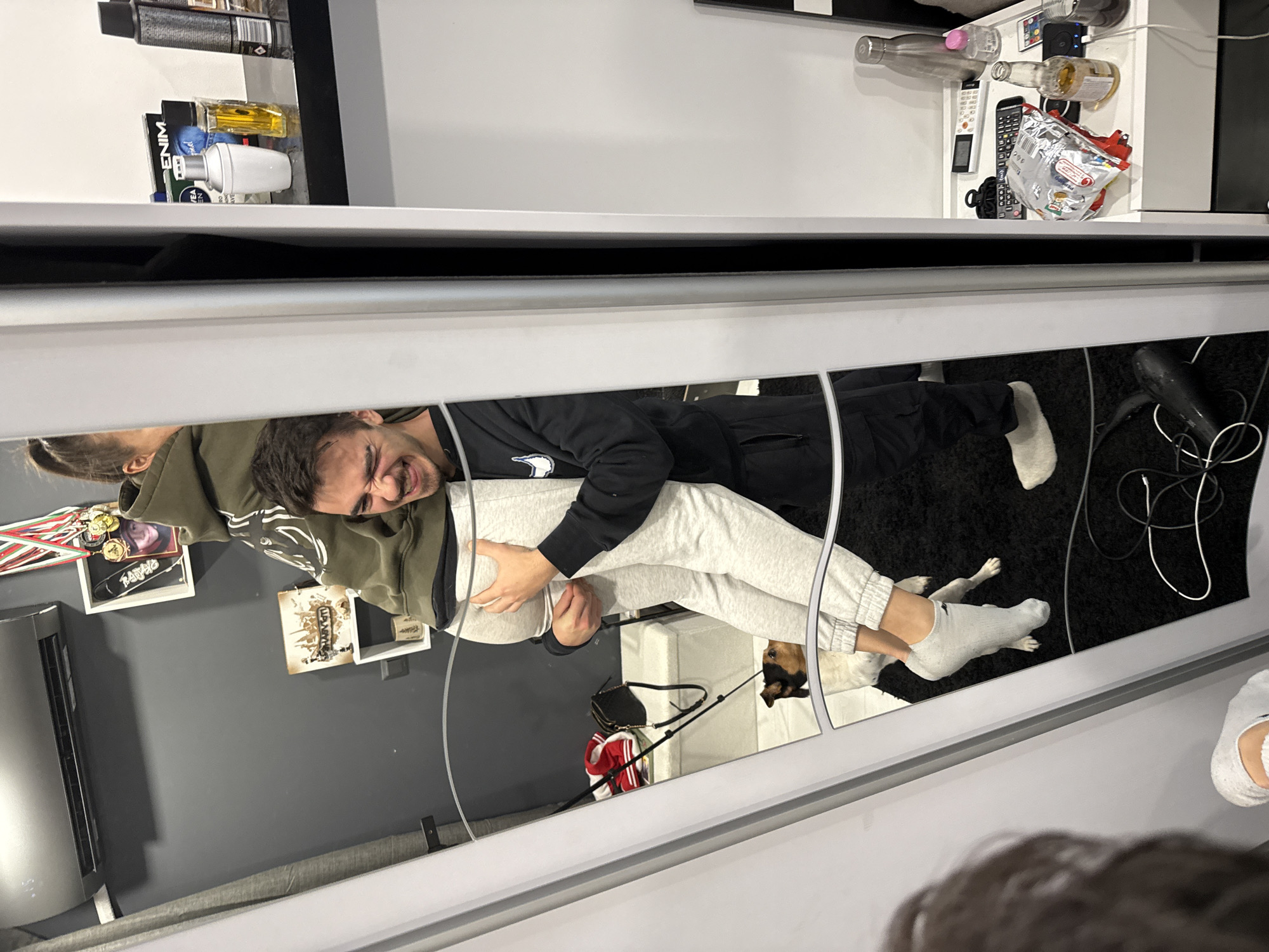 A sideways view shows a reflection in a mirror, with a man wearing a black tracksuit lying on a woman wearing a green hoodie and grey sweatpants. The reflection shows the woman holding a man around the torso. A dog lies on the floor by the legs of the sweatpants-wearing individual. Various items and objects are visible on shelves behind the mirror and in the mirrored reflection of the room, including framed items, awards, electronic devices, and bags. The exact location in Sofia, Bulgaria, cannot be determined from the image.