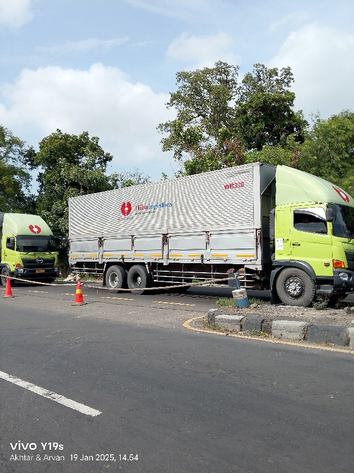 Two light-green cabover trucks, identified by "tibalogistics" branding and logo, are parked or positioned on the side of an asphalt road in Arjawinangun, Indonesia. The larger truck, on the right, features a long white box trailer marked "WB390" and has its front right wheel close to or on a concrete curb. A smaller "HINO" truck is visible further down the road on the left.
The scene is outdoors under a bright blue sky with scattered white clouds, suggesting a clear day. Two orange traffic cones are placed on the road between the trucks, connected by a yellow and black striped rope or barrier, indicating a temporary obstruction, warning, or work zone. Lush green trees line the background and sides of the road. The road itself has yellow and white markings.
No people are visible in the image. The trucks appear to be commercial vehicles, likely engaged in logistics or delivery services. The image includes a watermark stating "vivo Y19s", "Akhtar & Arvan", and a timestamp of "19 Jan 2025, 14.54".
