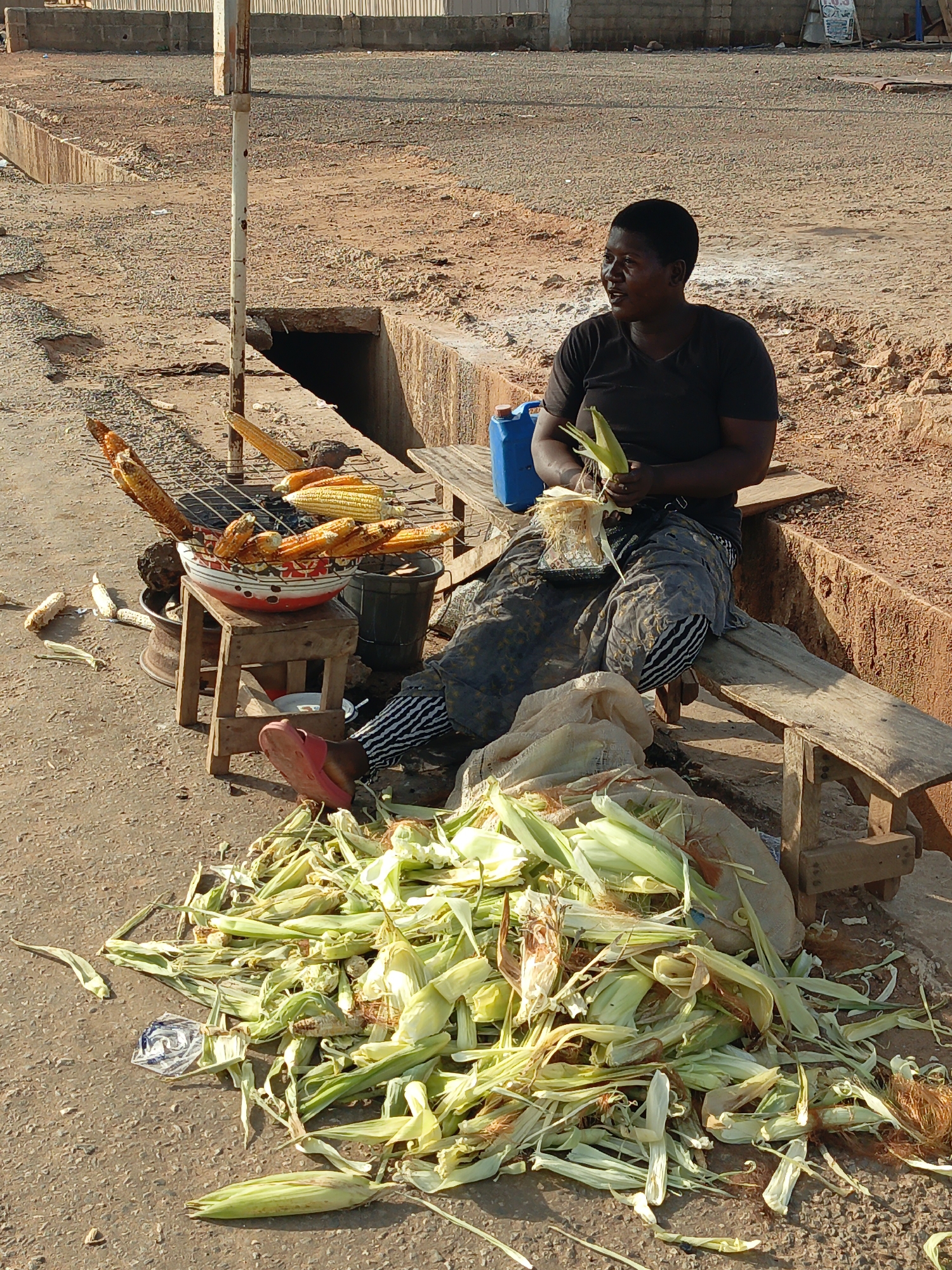 In Akwanga, Nigeria, a woman sits on a weathered wooden bench along a concrete drainage ditch, shucking corn. She wears a black t-shirt, and a patterned skirt, and pink sandals. A blue jerrycan sits beside her on the bench. A pile of corn husks lies on the ground. A small wooden stool beside her holds a red and white enamel bowl filled with grilled corn. A metal grill stands above the bowl. Several more grilled corn cobs lean against the bowl, and some sit directly on the ground.