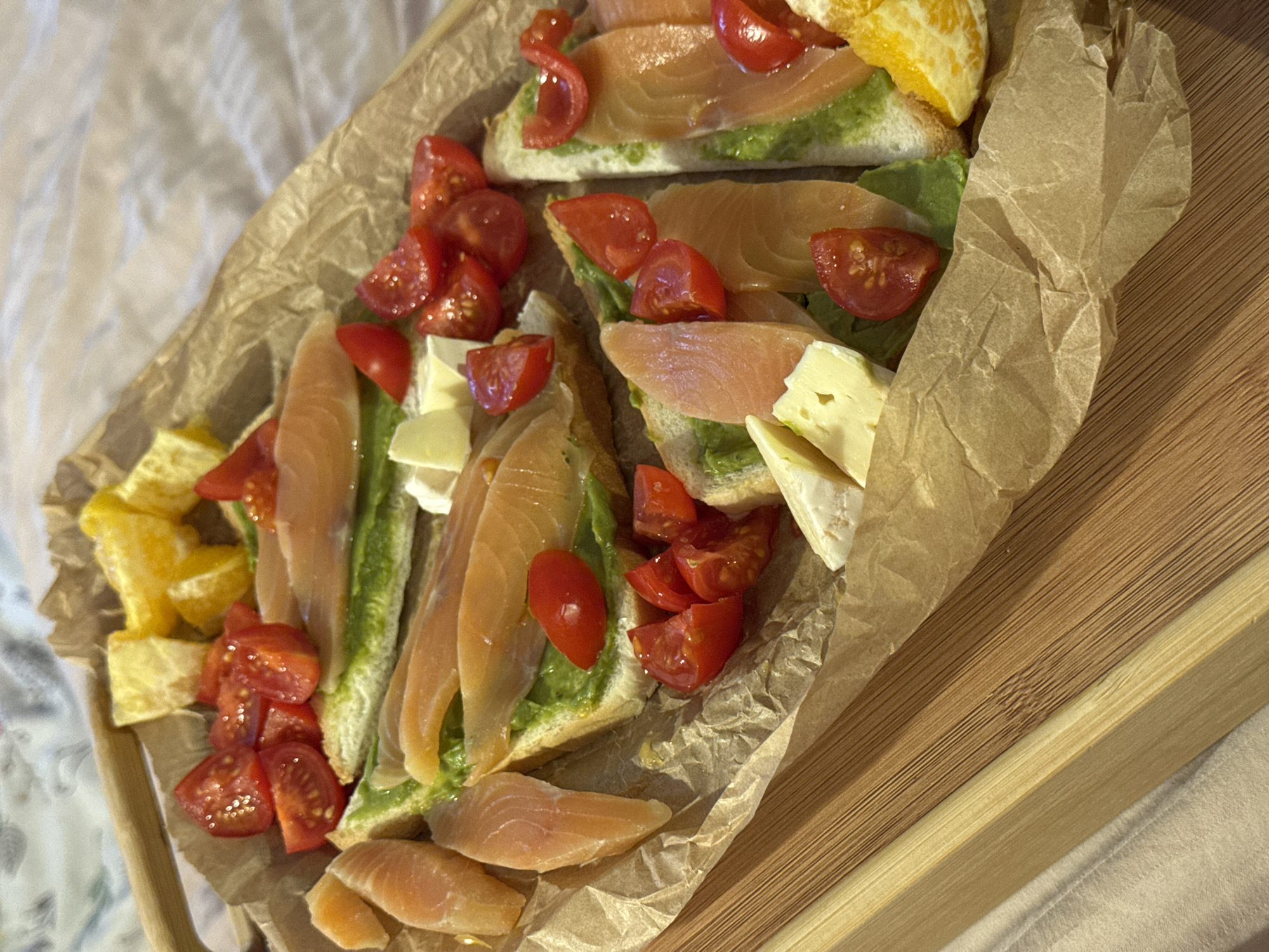 The image depicts an assortment of food items arranged on a piece of crinkled brown paper atop a wooden tray. The food includes open-faced sandwiches with a green spread, topped with slices of salmon, halved cherry tomatoes, and chunks of cheese and orange. The tray appears to be sitting on a light-colored textured surface, possibly a bedspread. The location is unknown, and there is no information to suggest that the photo was taken in Chaikovsky, Russia.
