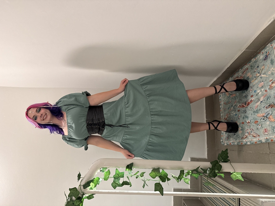A woman with pink and purple hair and fairy accessories smiles while posing in a green dress with a black corset. She stands on a tiled floor with a rug. Her attire includes black ballet flats with crossed straps. She leans against a white handrail adorned with artificial ivy leaves, giving the image a fairytale or whimsical aesthetic. The setting appears to be a hallway or staircase.