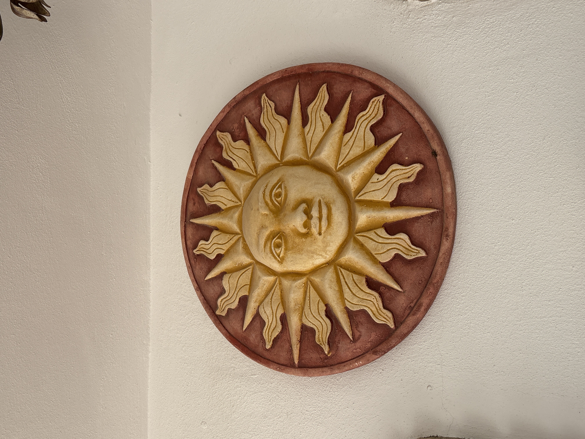 An ornate, circular sunburst motif, rendered in a textured, ceramic-like material, is affixed to a textured, off-white wall. The central element of the motif is a stylized human face with closed eyes and a subtle smile, painted in a golden yellow hue. Emanating from the face are numerous rays of light, also in golden yellow, each featuring a flame-like, undulating pattern. The outer border of the sunburst is a reddish-brown, circular disc that frames the golden rays. The wall itself is a pale, neutral color with a rough, stucco-like finish. The lighting appears to be natural, casting subtle shadows that highlight the relief of the sunburst. The image is tightly cropped, focusing solely on the decorative piece and its immediate surroundings, with no indication of the broader environment or specific location within Orihuela, Spain.