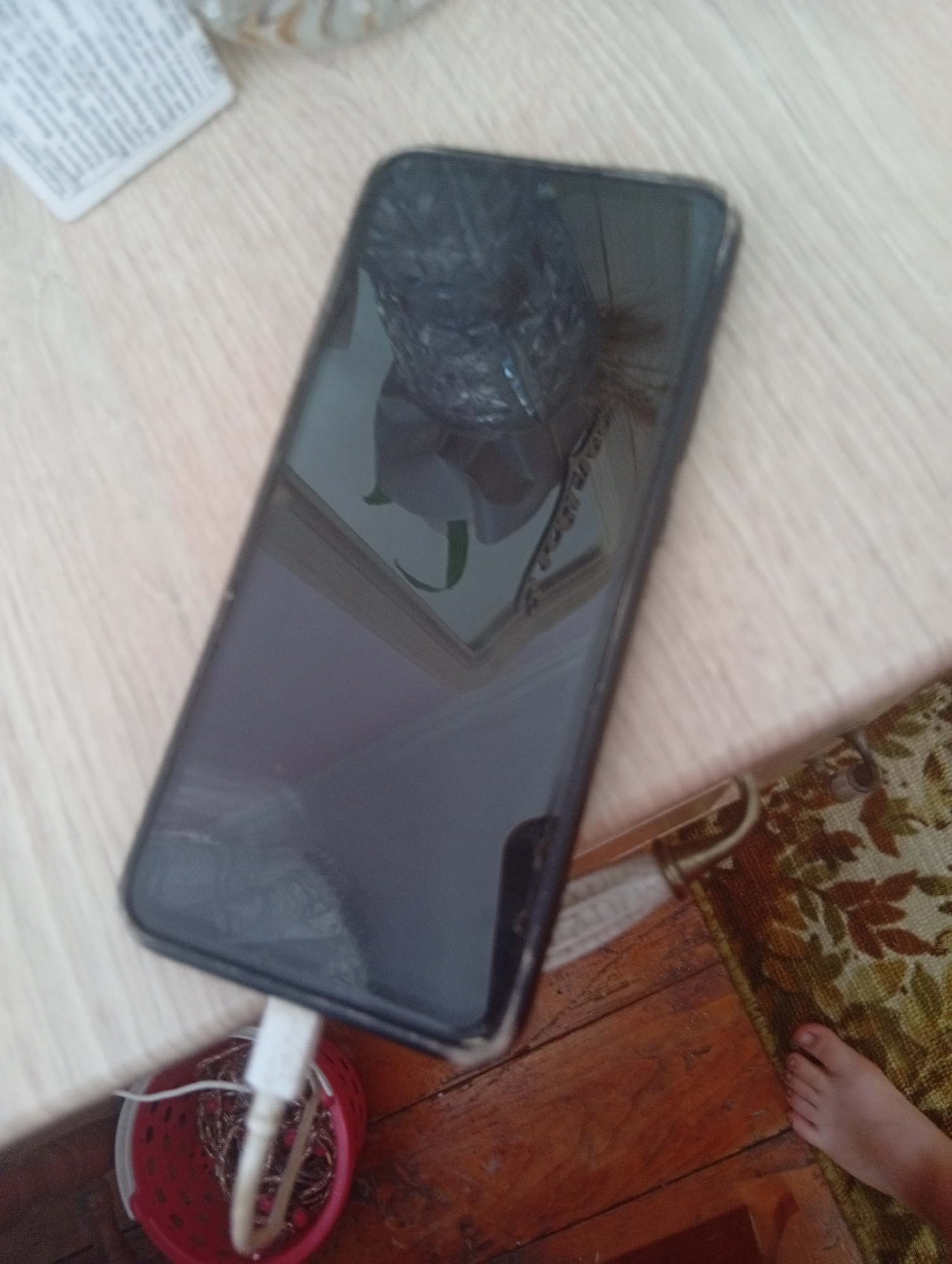 A black smartphone is lying on a wooden table. The screen of the phone is cracked and reflects a distorted image of the surroundings. A white charging cable is plugged into the bottom of the phone and goes into a small red container filled with what appears to be wires or necklaces. The table has a light-colored wood grain and is next to a wooden floor. A rug with a floral pattern is partially visible on the right side of the image, with a bare foot resting on it. The scene appears to be indoors. This scene is located in Veliatyno, Ukraine.