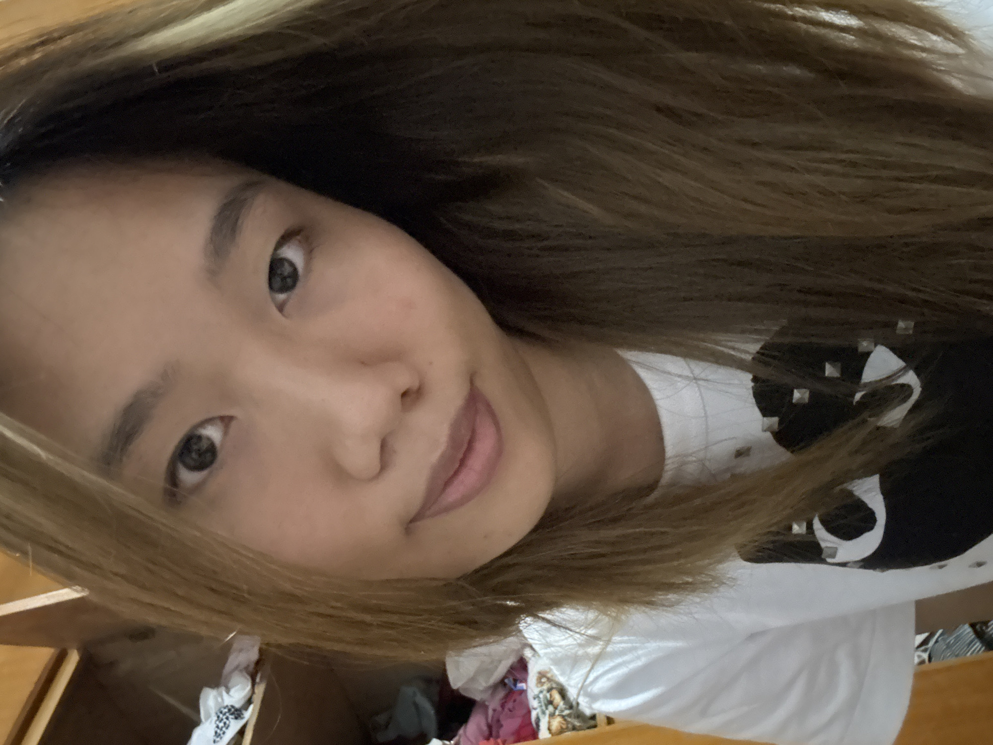 A close-up, head-and-shoulders selfie shows a young East Asian woman with medium-length, light brown hair with blonde highlights. She is wearing a white t-shirt with a black graphic design featuring a heart shape composed of smaller heart shapes, studded with silver accents. Her skin is light-toned, and she has dark eyes and eyebrows. The background is dimly lit and appears to be an indoor domestic setting, possibly a bedroom or closet, with wooden shelving and some clothing visible. The image is not geographically specific to Guandu, China.