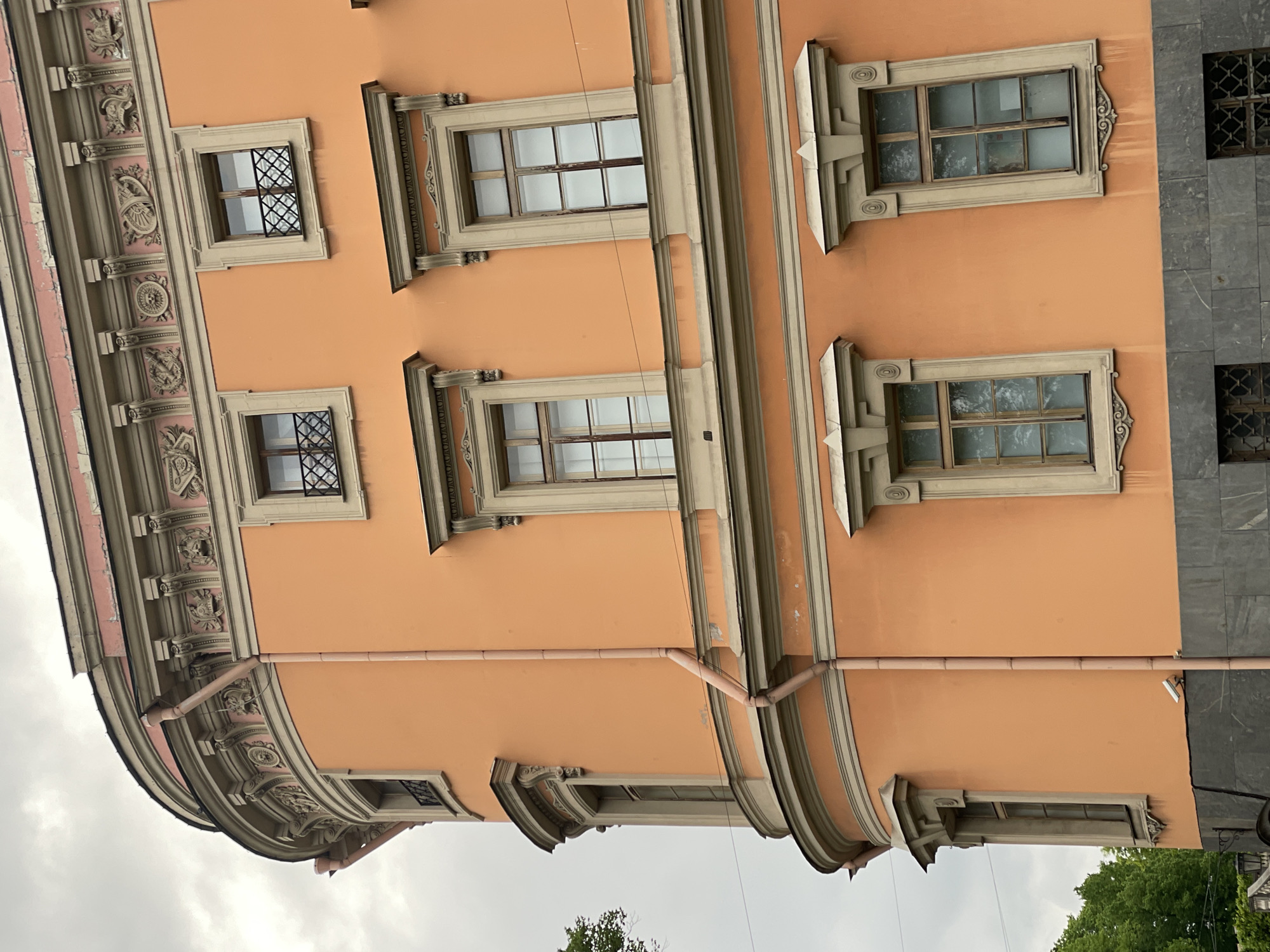 The image depicts a section of a building in Saint Petersburg, Russia. The facade is a peach color with decorative architectural elements, including ornate window frames and sculpted details along the building's edge. Various windows are visible, some with lattice-like grilles. A drainage pipe runs down the side of the building. The sky is overcast, and the top of a tree is visible in the lower right corner.