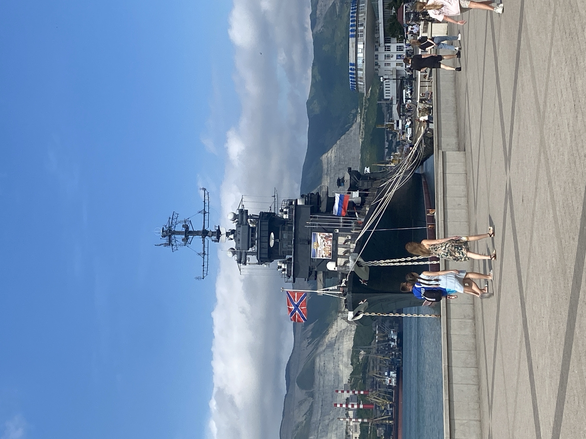 A large warship is docked at a harbor in Novorossiysk, Russia, with its bow facing the viewer. A Russian naval ensign flies from a mast on the ship's deck. A banner depicting a group of people is also visible on the ship. In the foreground, a paved walkway runs along the harbor, with several people walking on it. The people are dressed in casual summer clothing. In the background, there are industrial buildings, cranes, and other ships in the harbor. Mountains rise behind the city, and the sky is blue with some clouds.