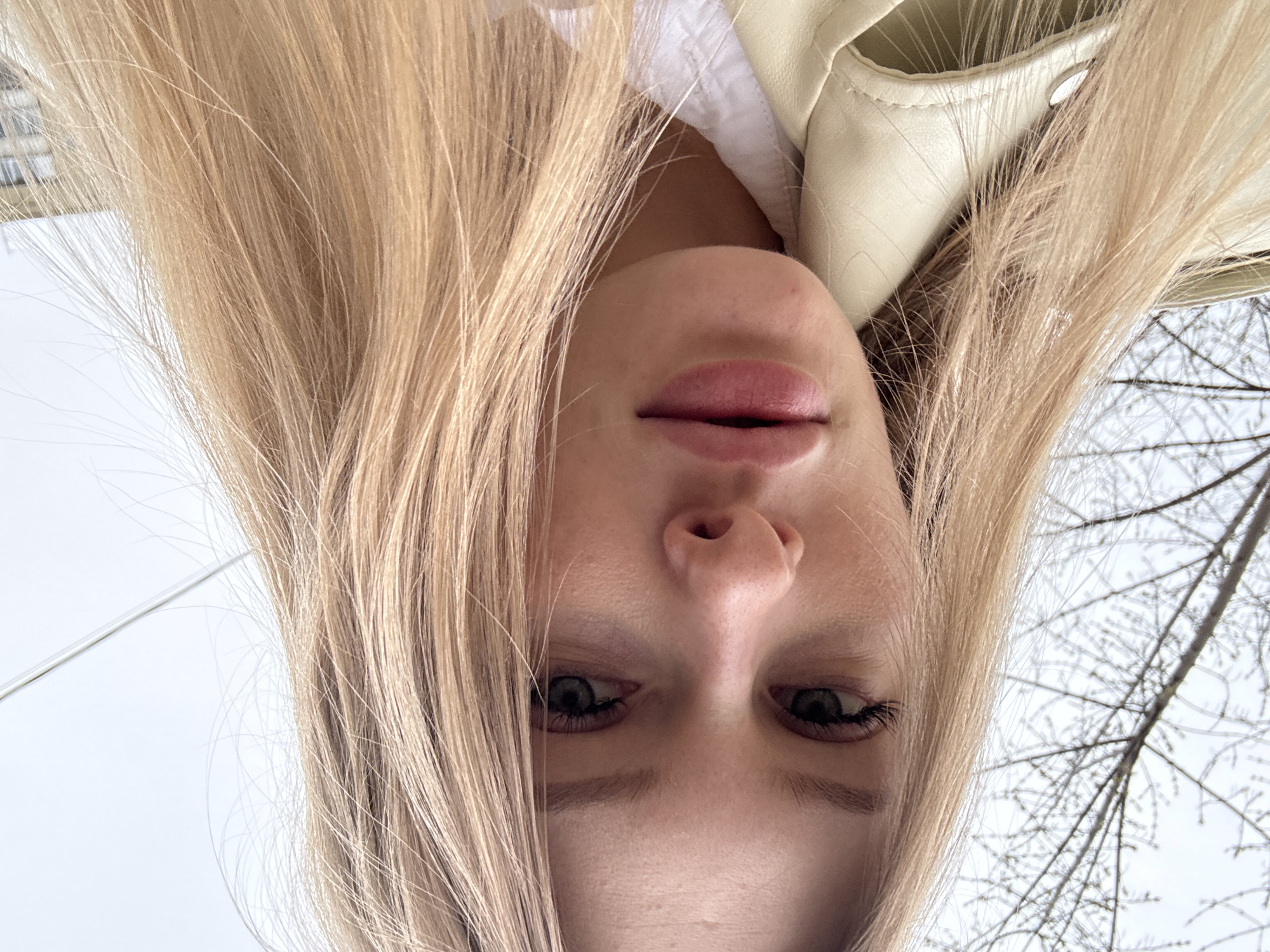 A close-up selfie of a woman with long blonde hair, taken at an upward angle. Her face is centered in the frame, with her eyes, nose, and mouth visible. She appears to be wearing a white top and a light-colored leather jacket. The background includes a gray sky and bare tree branches. In the upper left corner, a building is faintly visible. Based on the general architectural style and the presence of bare trees in an urban setting, it is plausible that the photo was taken in a city like Perm, Russia, though this cannot be definitively confirmed from the image alone.