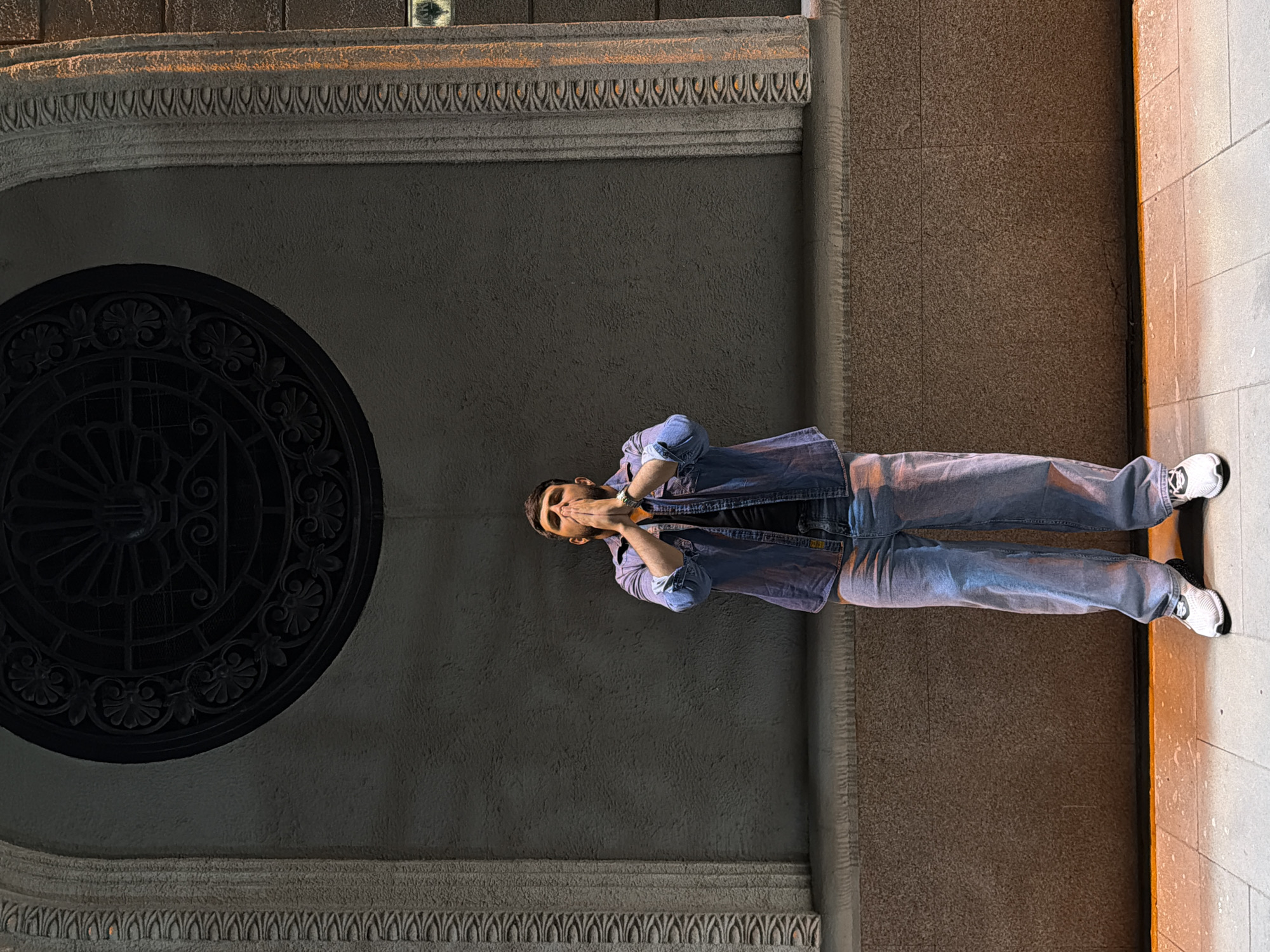 A full-length shot captures a man standing within an archway in Krasnoyarsk, Russia. The man is wearing a light-blue denim jacket over a black t-shirt, blue jeans, and white athletic shoes. His hands are raised to his face, as if in a gesture of surprise or deep thought.
The archway features a circular ornamental design on the upper left, and decorative molding along the top. The right side of the archway is a light-brown stone-tiled wall. The image is rotated 90 degrees clockwise.