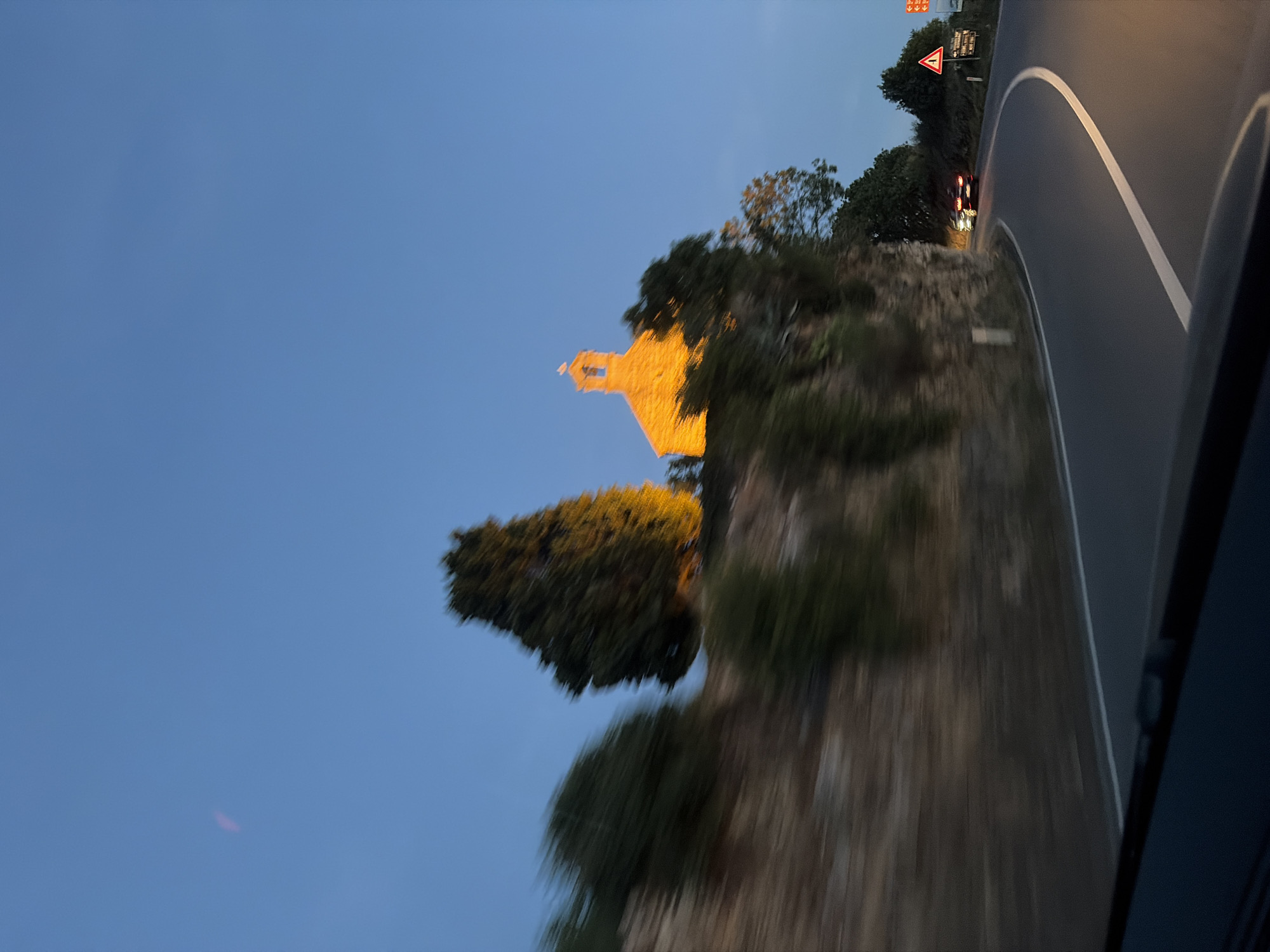 The image depicts a winding road at dusk, likely in or near Blizikuće, Montenegro. A small, stone building, appearing to be a church, is illuminated by artificial lights on a hill overlooking the road. The sky is a deep blue, transitioning from daylight to night. The road curves to the right, with a white line marking its edge. Two cars are visible on the road, one in the distance with its brake lights illuminated. The surrounding landscape is characterized by a rocky slope and trees, some of which are blurred due to motion. Road signs are also present, indicating potential hazards or directions.