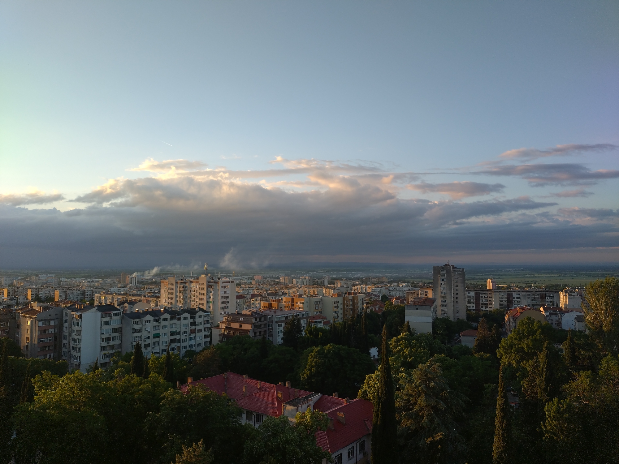 The image captures a high-angle view of the city of Stara Zagora, Bulgaria, during early morning or late evening. The foreground contains dense greenery consisting of various trees, including tall evergreens. Several buildings are visible, with red tiled roofs peeking through the foliage. Beyond the greenery is a cityscape of predominantly multi-story residential buildings, exhibiting a range of architectural styles. Some buildings feature white facades and visible balconies. In the distance, a flat plain stretches towards the horizon. The sky is a gradient of light blue and soft pink hues, with a layer of clouds partially covering the horizon. Some buildings are emitting smoke.
